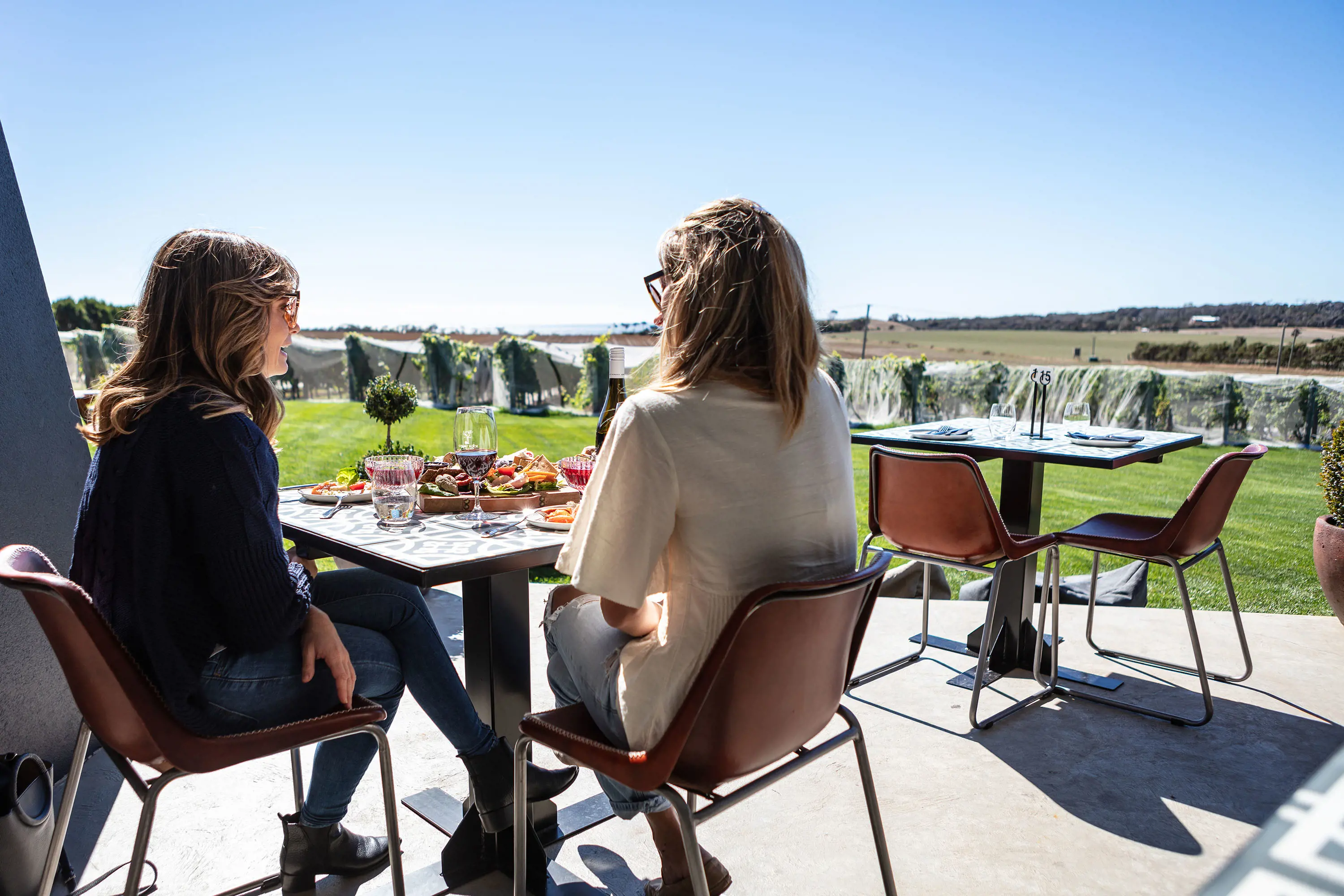 Two women seated outdoors at a table full of food and wine by a vineyard.