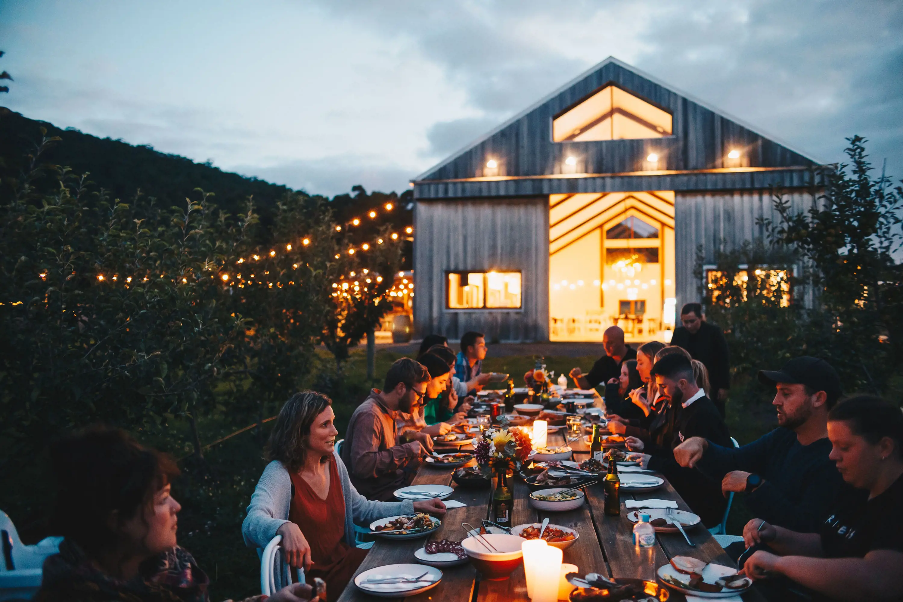 Outside at twilight, in front of a large lit-up barn, people sit dining along a long table filled with plates of food and drinks.