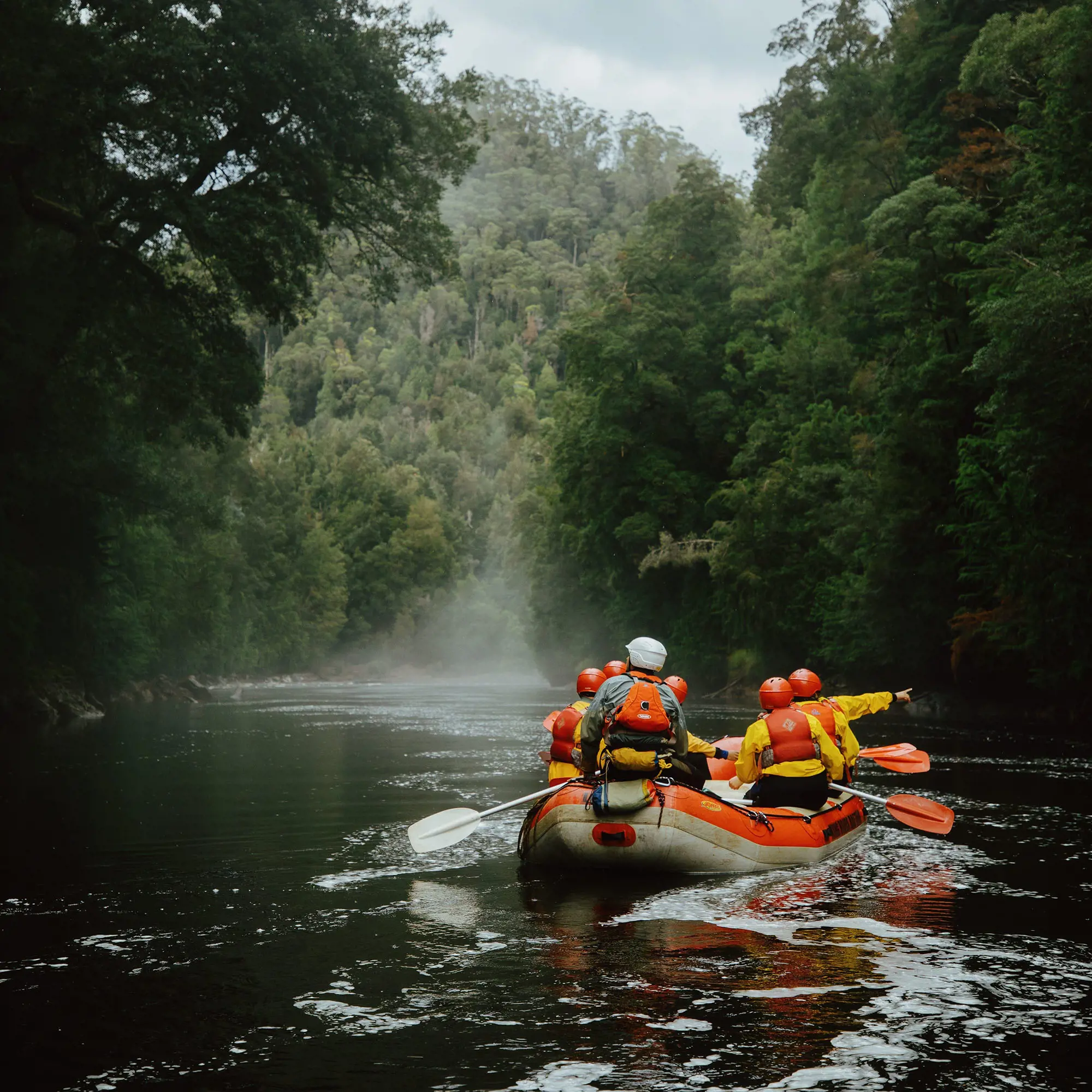 People in orange lifejackets and helmets paddle an inflatable dinghy down a misty, atmospheric river, bordered by tall lush forest.