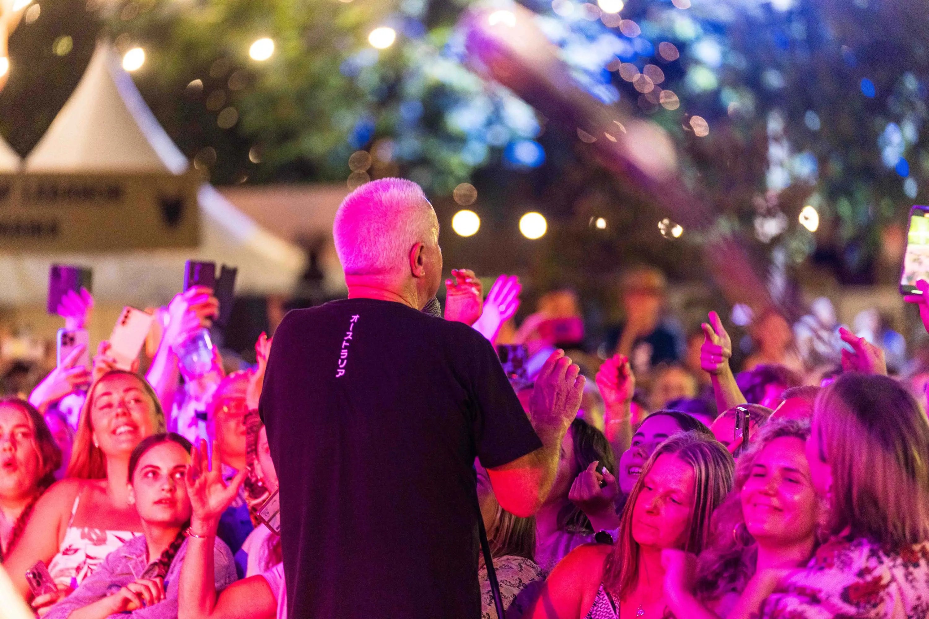 A man sings to a crowd of people bathed in a pink light, with their hands in the air and dancing.