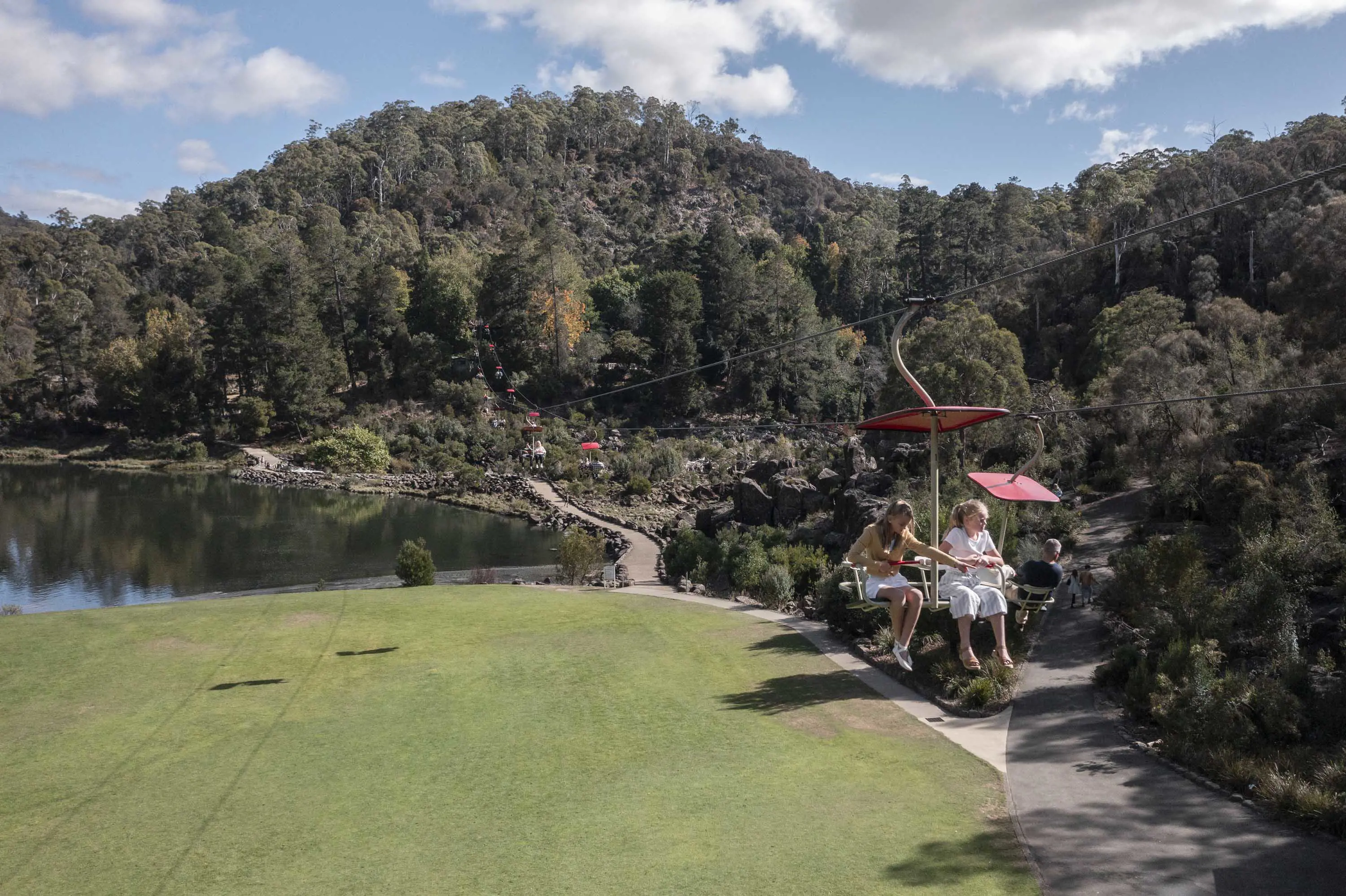 Two girls ride a chairlift stretched across parklands and large lake within a valley.