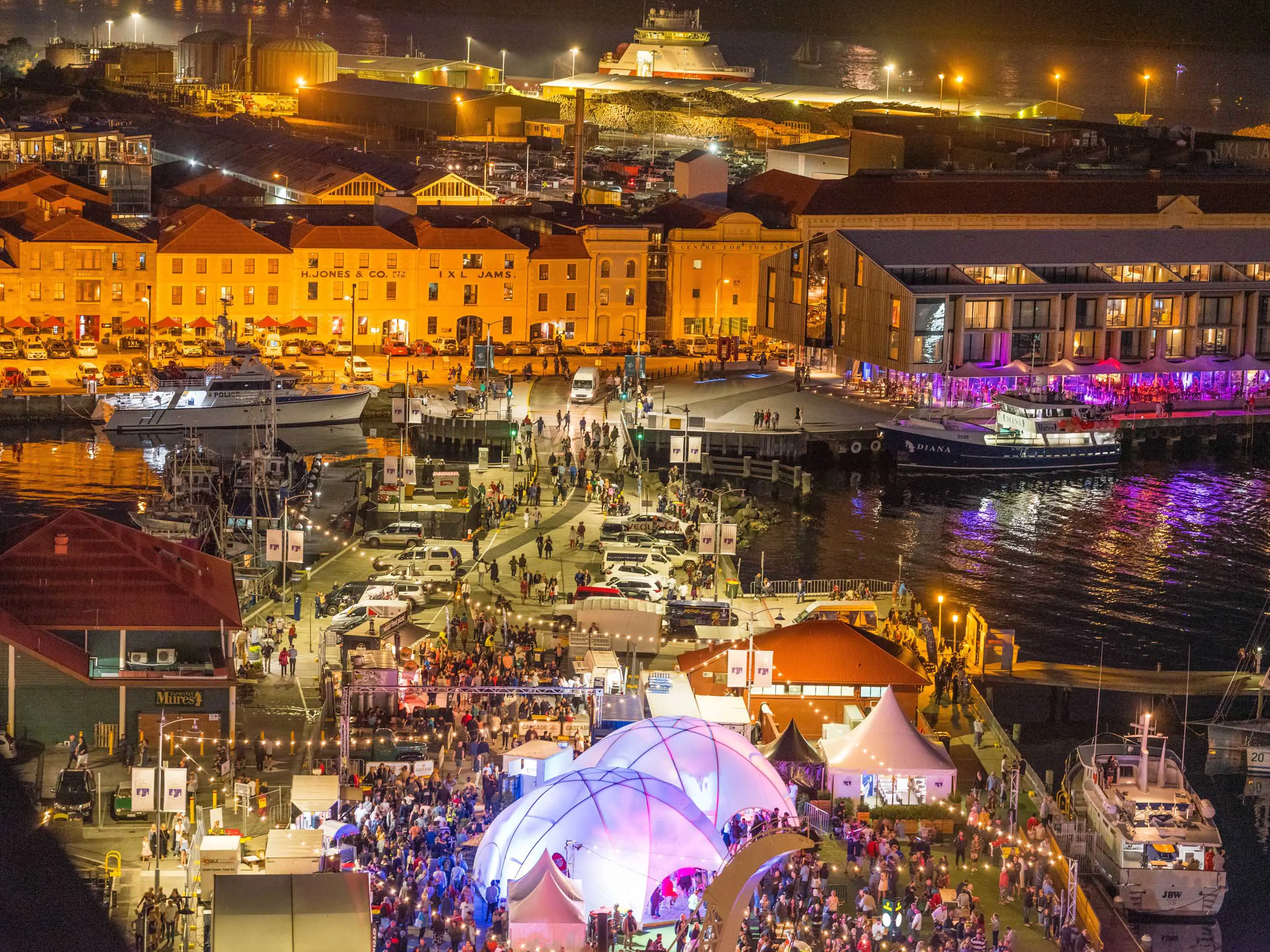 An aerial view of the Hobart waterfront with large crowds of people mingling amongst tents and stalls.