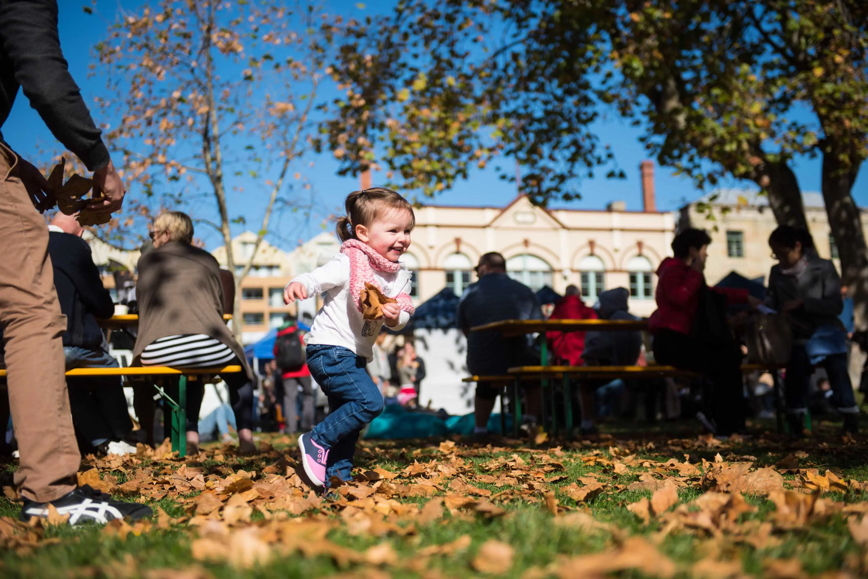 A small child runs in autumn leaves on the grass in front of stalls and Georgian style buildings in a sunny day.