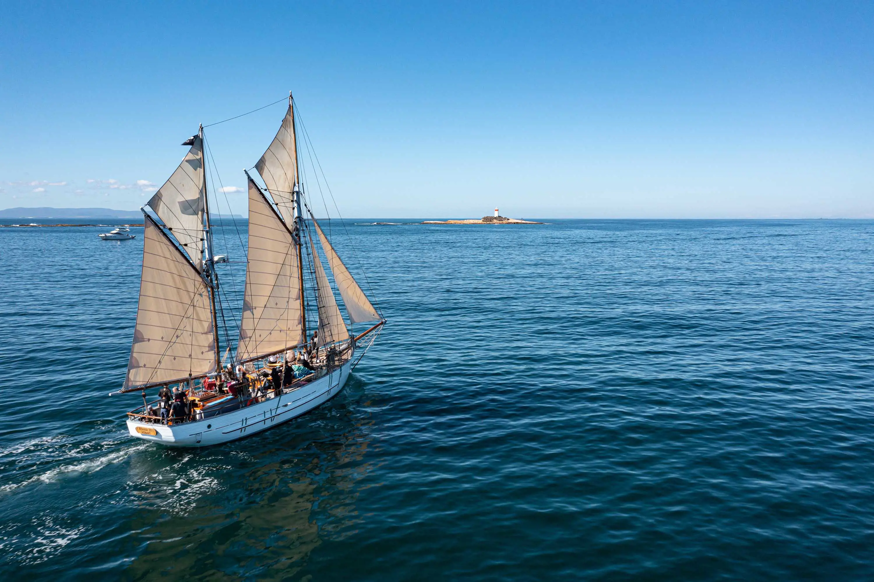 A large, two-mast wooden boat with sails glides across calm waters on a clear, sunny day.