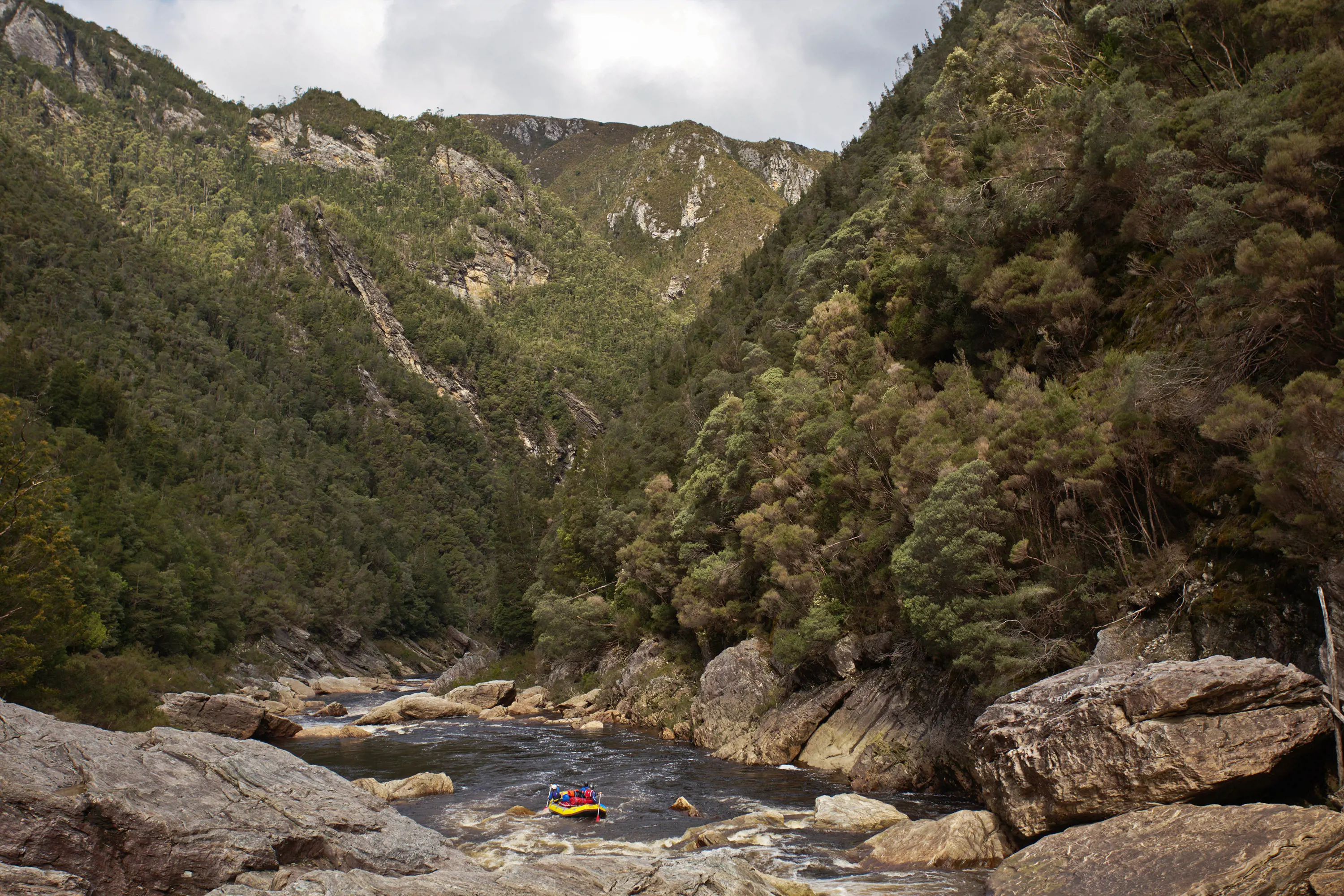 People rafting in a yellow inflatable boat are dwarfed by a wide, fast-moving rocky river. The landscape around rises up sharply and is densely bushy.