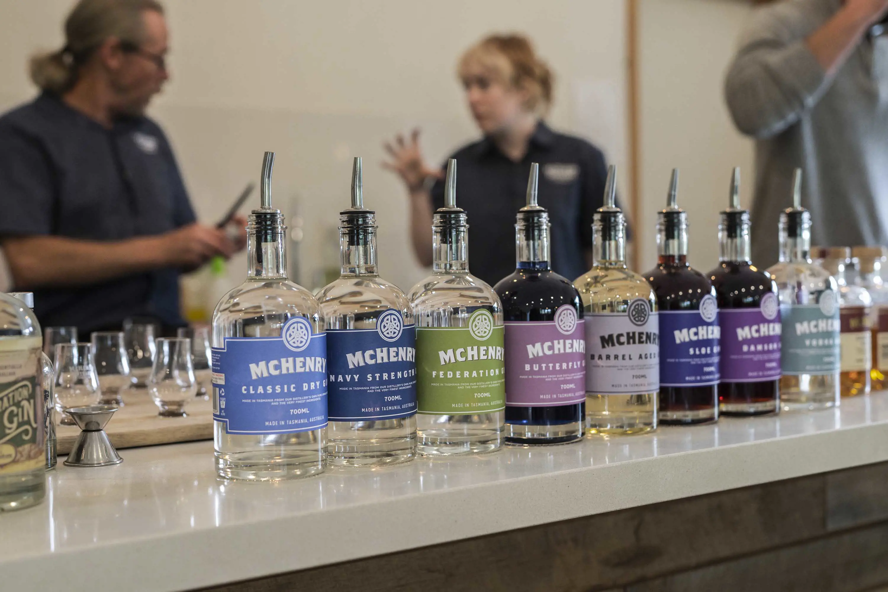 A selection of specialty gins are lined up on a solid stone counter in a tasting room. People talk in the background.