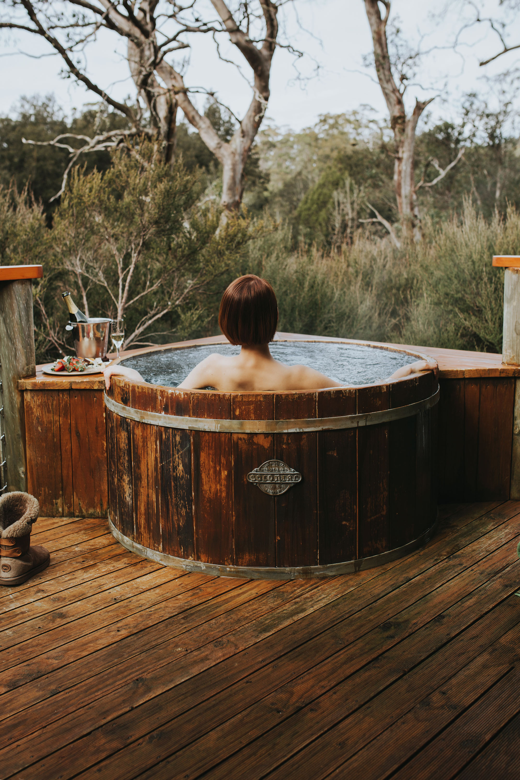 A woman soaks in an outdoor hot tub on a wooden deck looking out into the bush. Beside her sits champagne and a plate of strawberries.