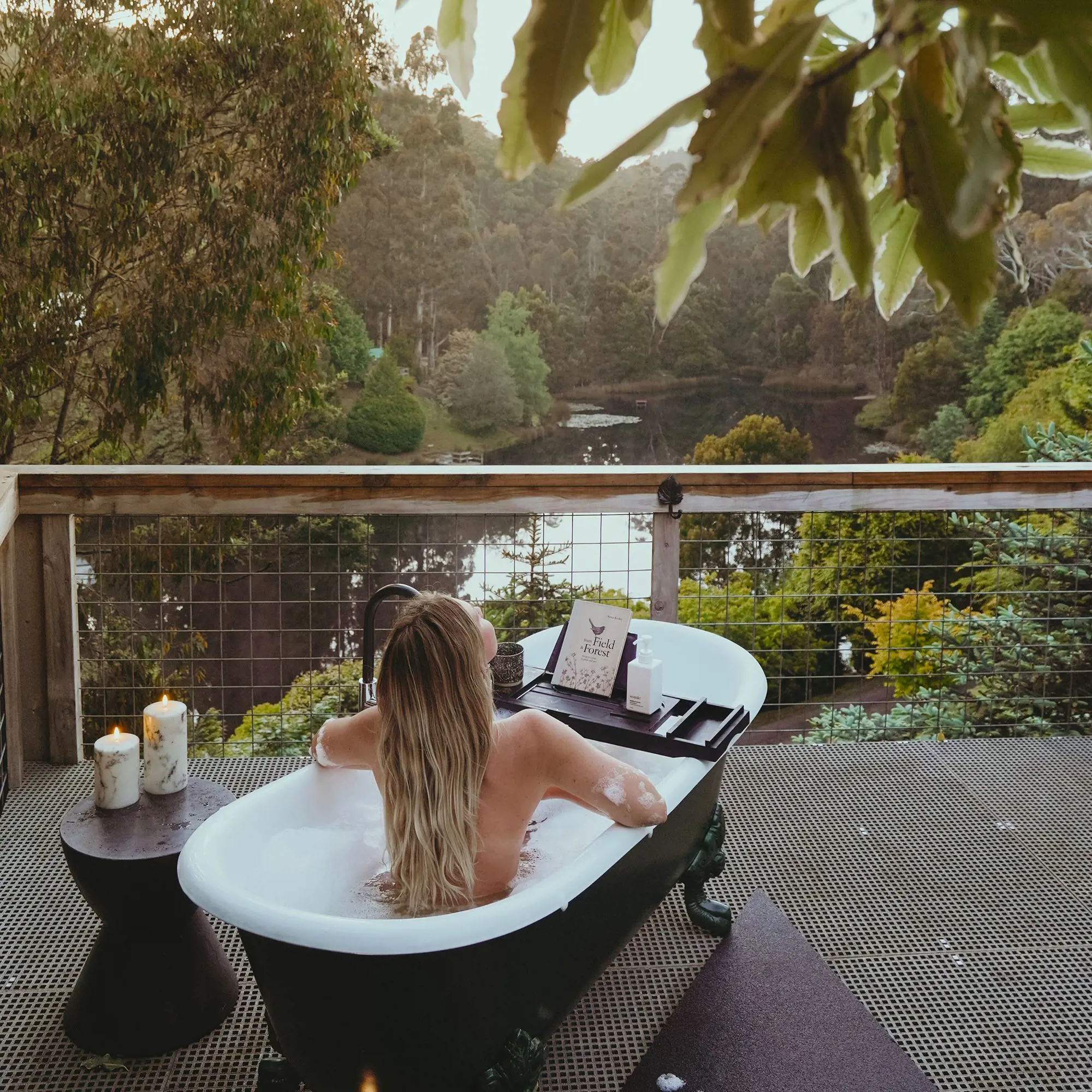 A woman relaxes in a black and white outdoor bathtub on a balcony overlooking the wilderness.