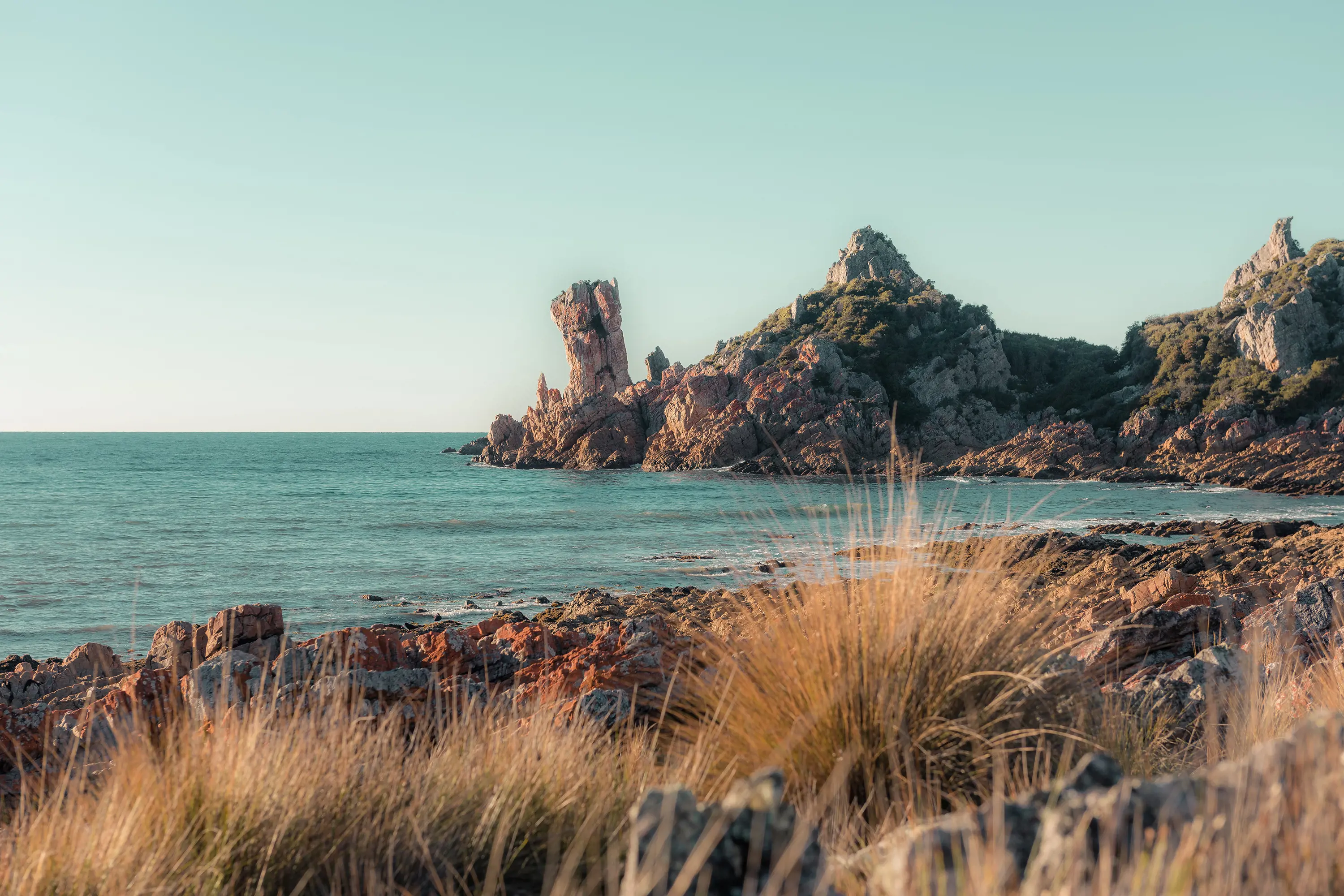 From a rocky, grassy beach, a view over the water to an unusual tall cylindrical rock formation jutting out of the rocky shoreline.