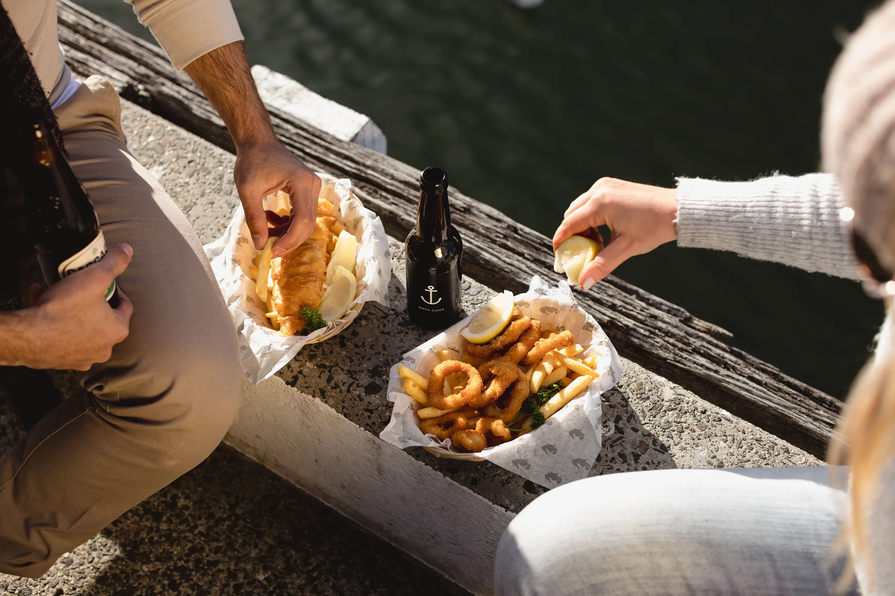 Two baskets of seafood and chips sit on the edge of a wharf, with the dark water below. People are midway through reaching for the food.