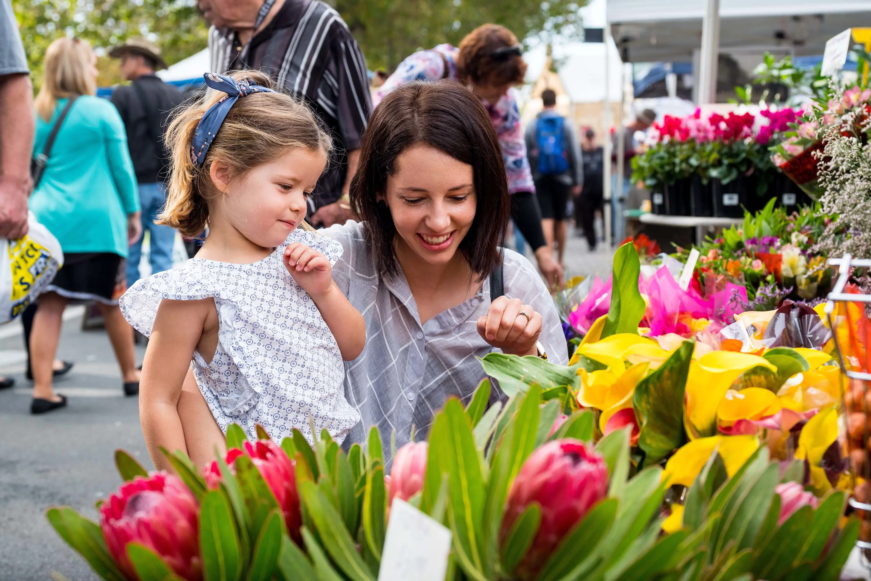 In a busy outdoor market, a mother and daughter look at colourful yellow and pink flowers with greenery.