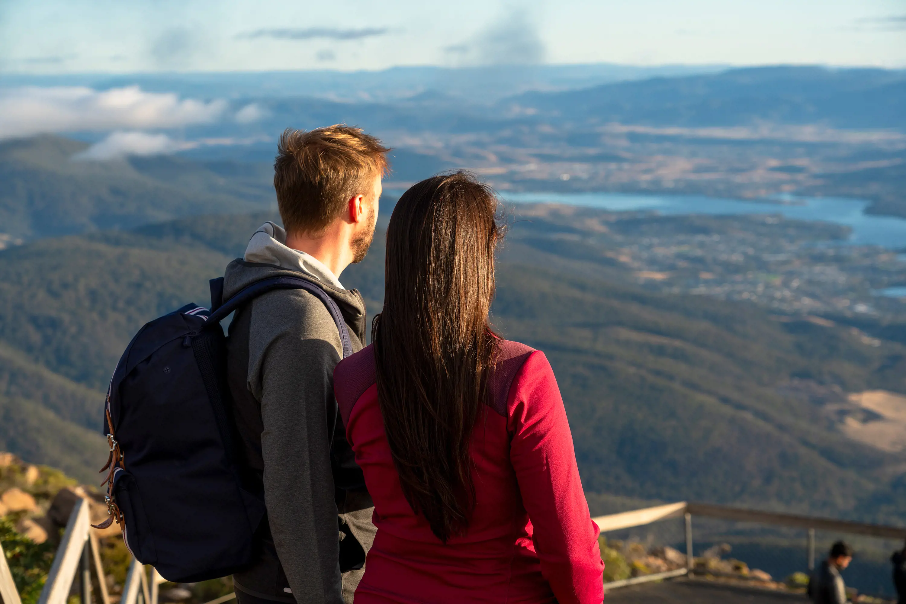 A man and a woman look out from a viewing platform atop a mountain, with a river snaking through a city in the distance.