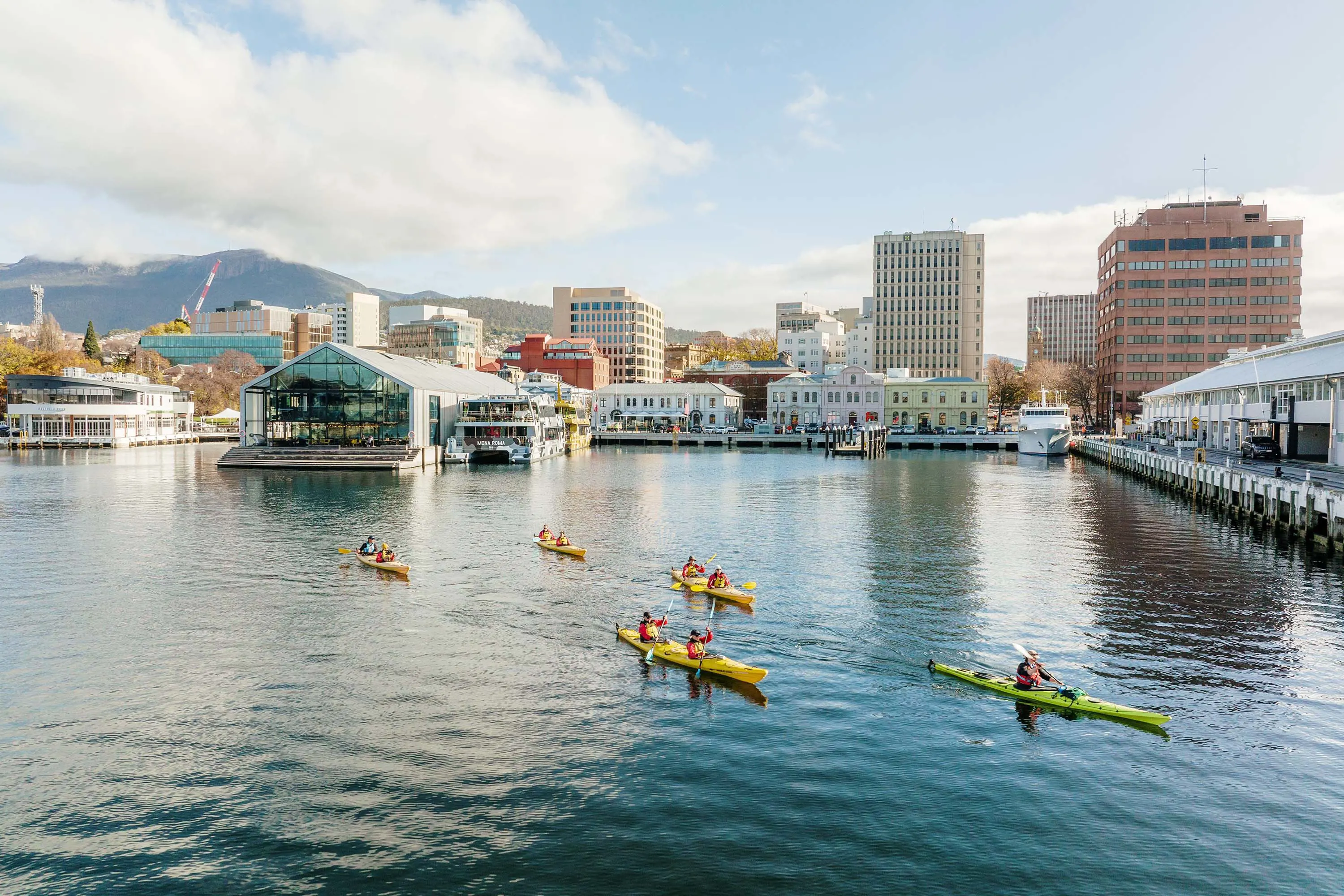 A group of kayakers in yellow and green kayaks paddle across the dappled river water of a city harbour.