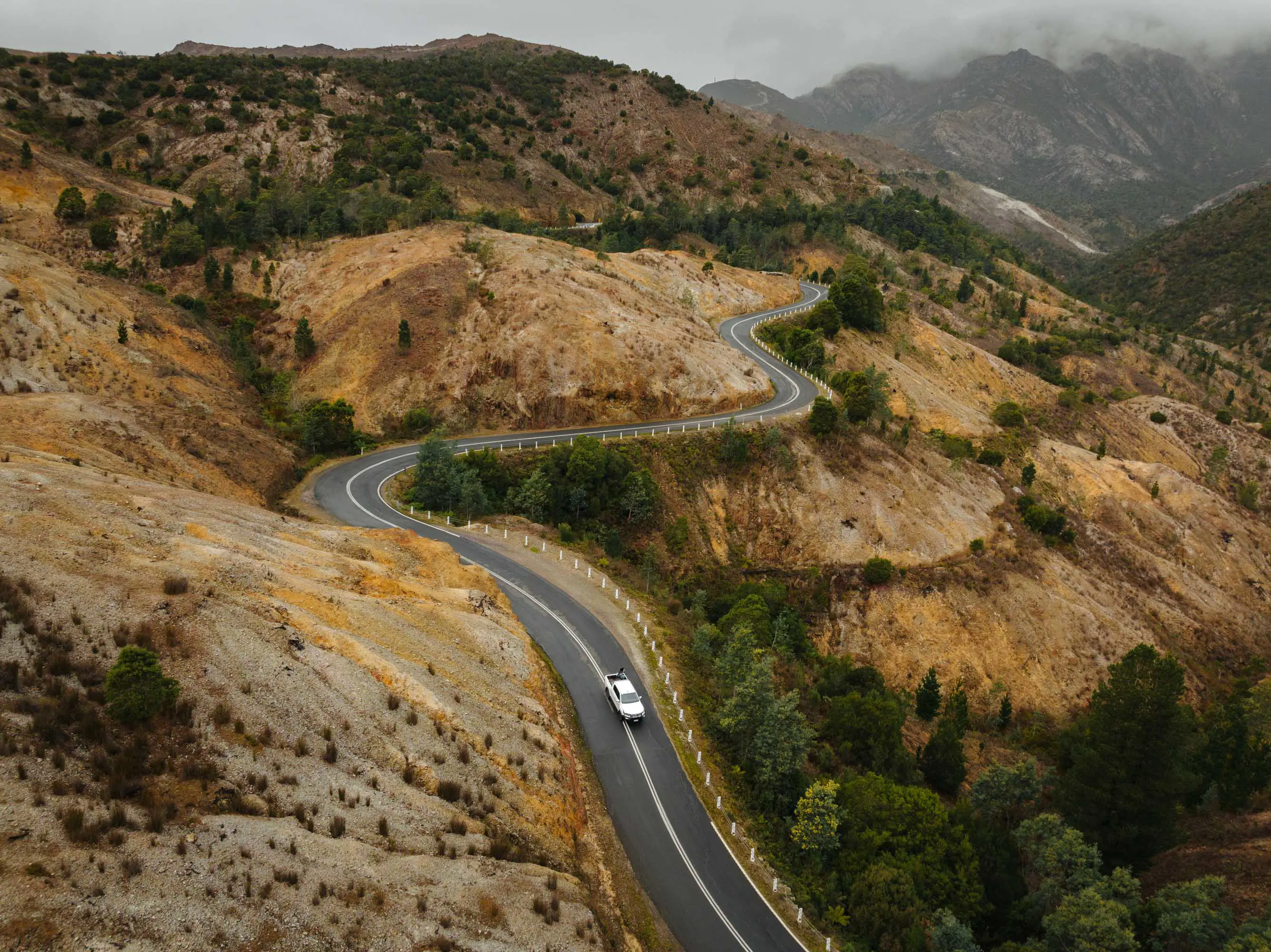 A white vehicle travels along a winding road through ochre-coloured mountain ranges on an overcast day.