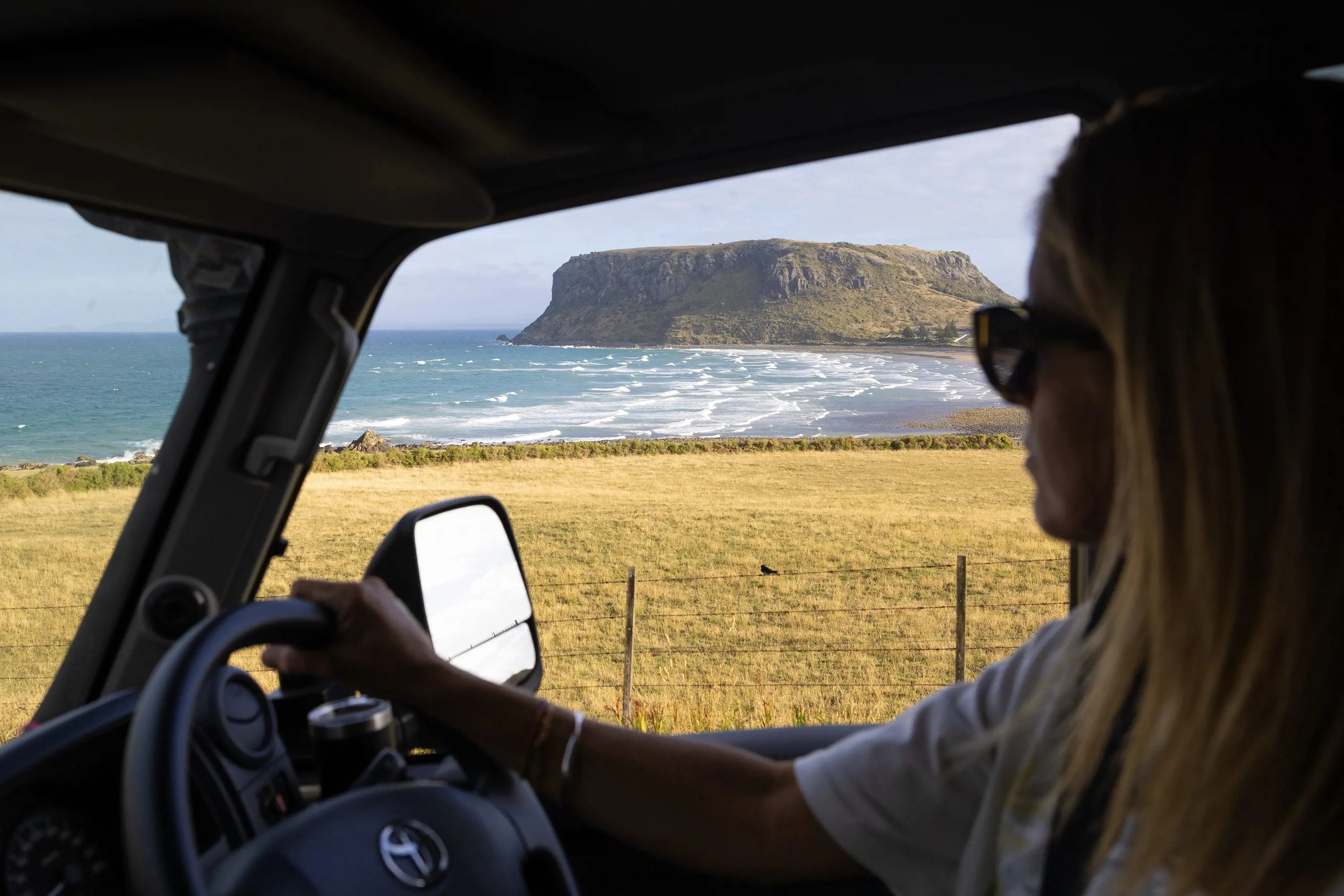 From the passenger seat of a car, looking past the driver out to the ocean and a large flat-topped hill.