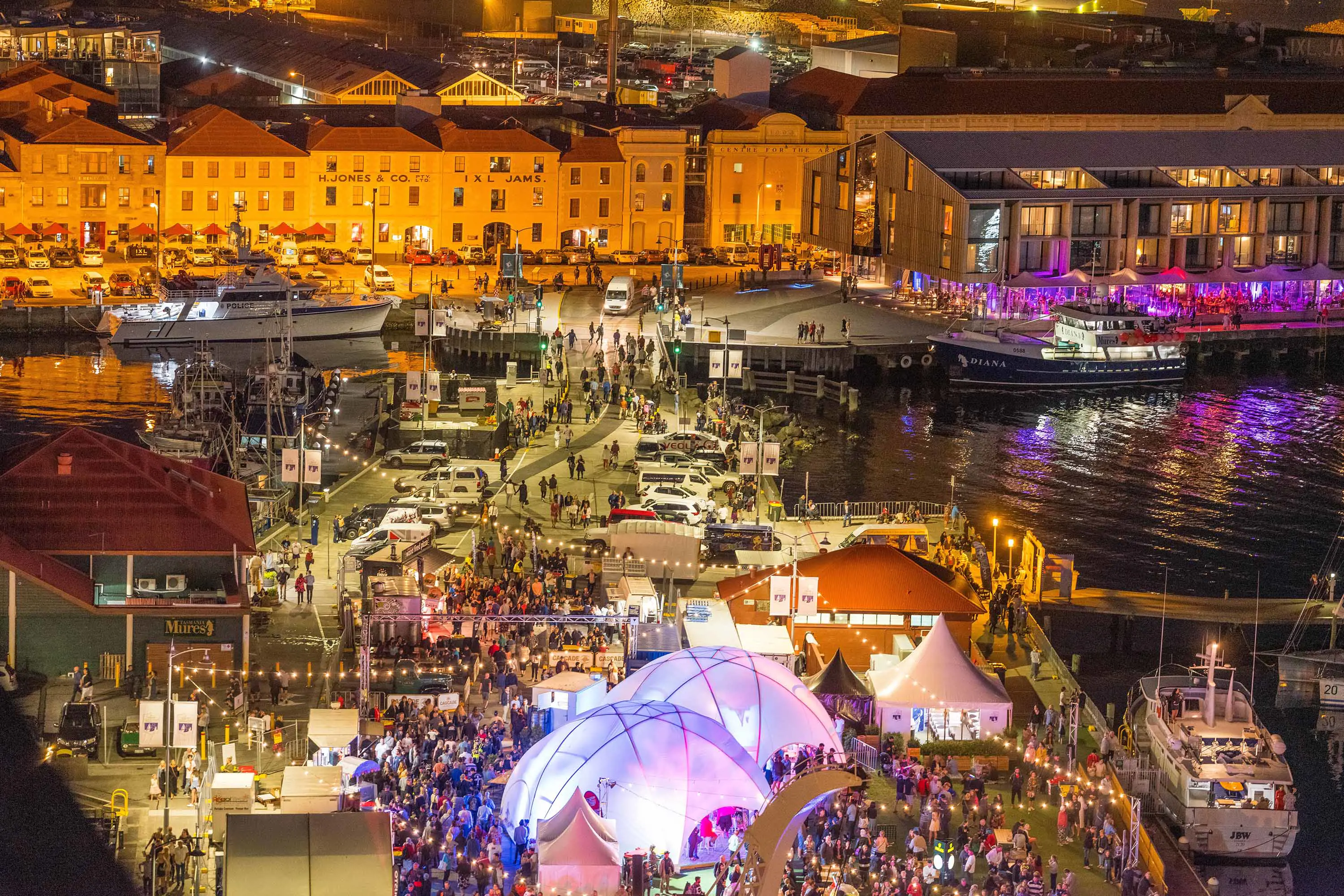 An aerial view of a busy waterfront event at night. People crowd in an area with colourful lights and marquees, while a facade of historic buildings glow brightly.