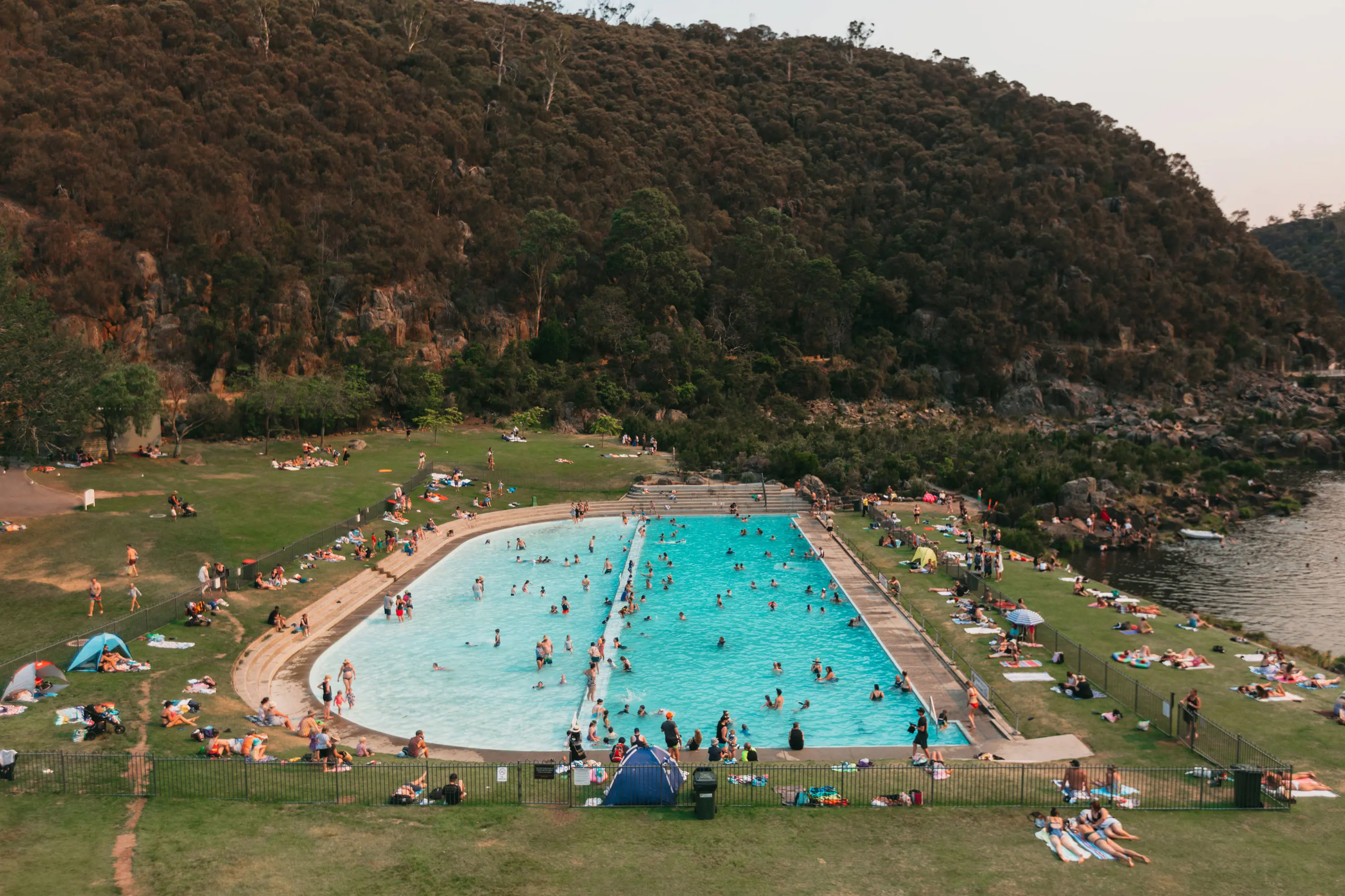 A large, semicircular aqua outdoor pool set into a grassy area next to a river, with lots of people swimming and relaxing. Behind is a steep, densely bushy cliff.