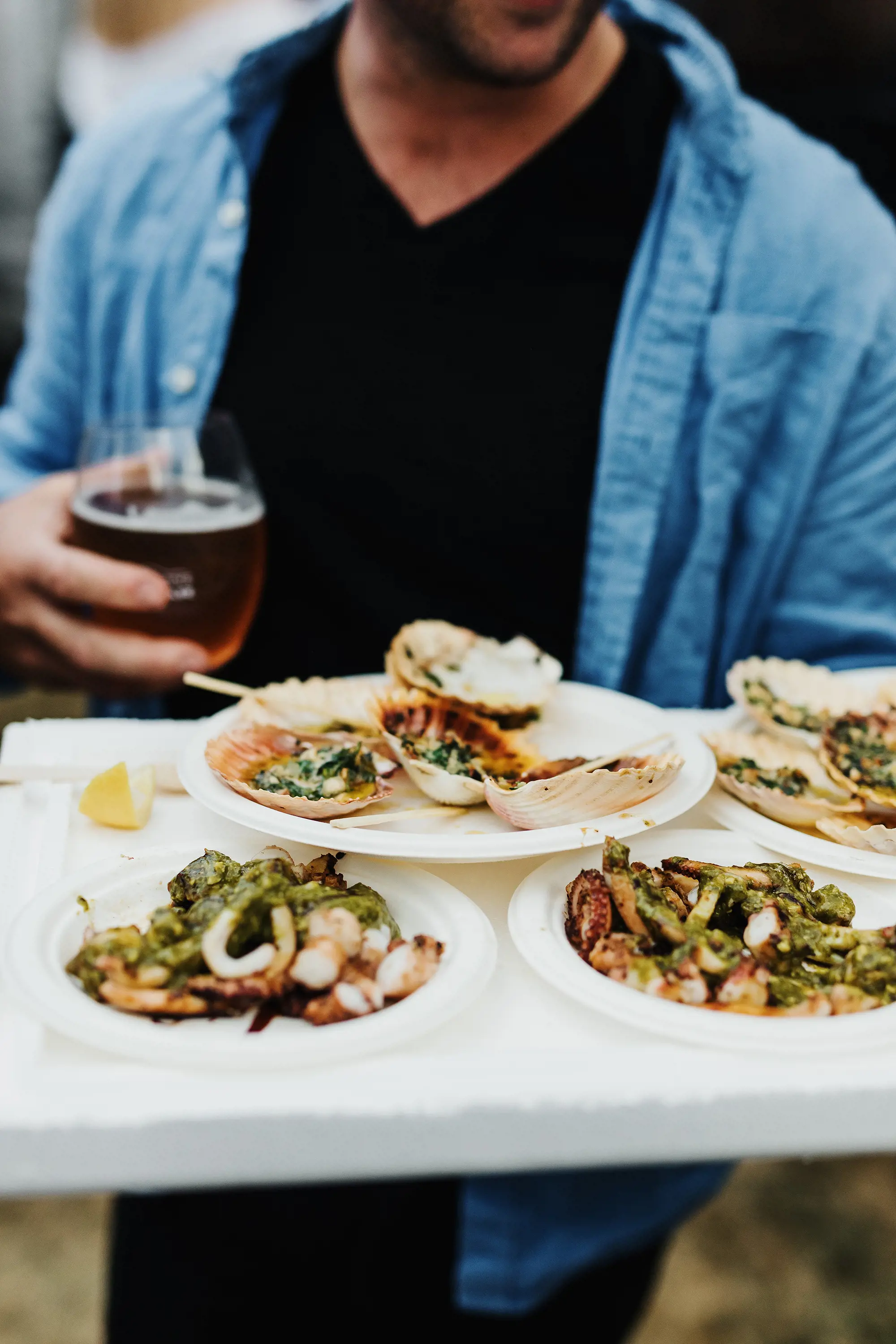 A person holding a tray with plates of cooked seafood in one hand, and a plastic cup of beer in the other.