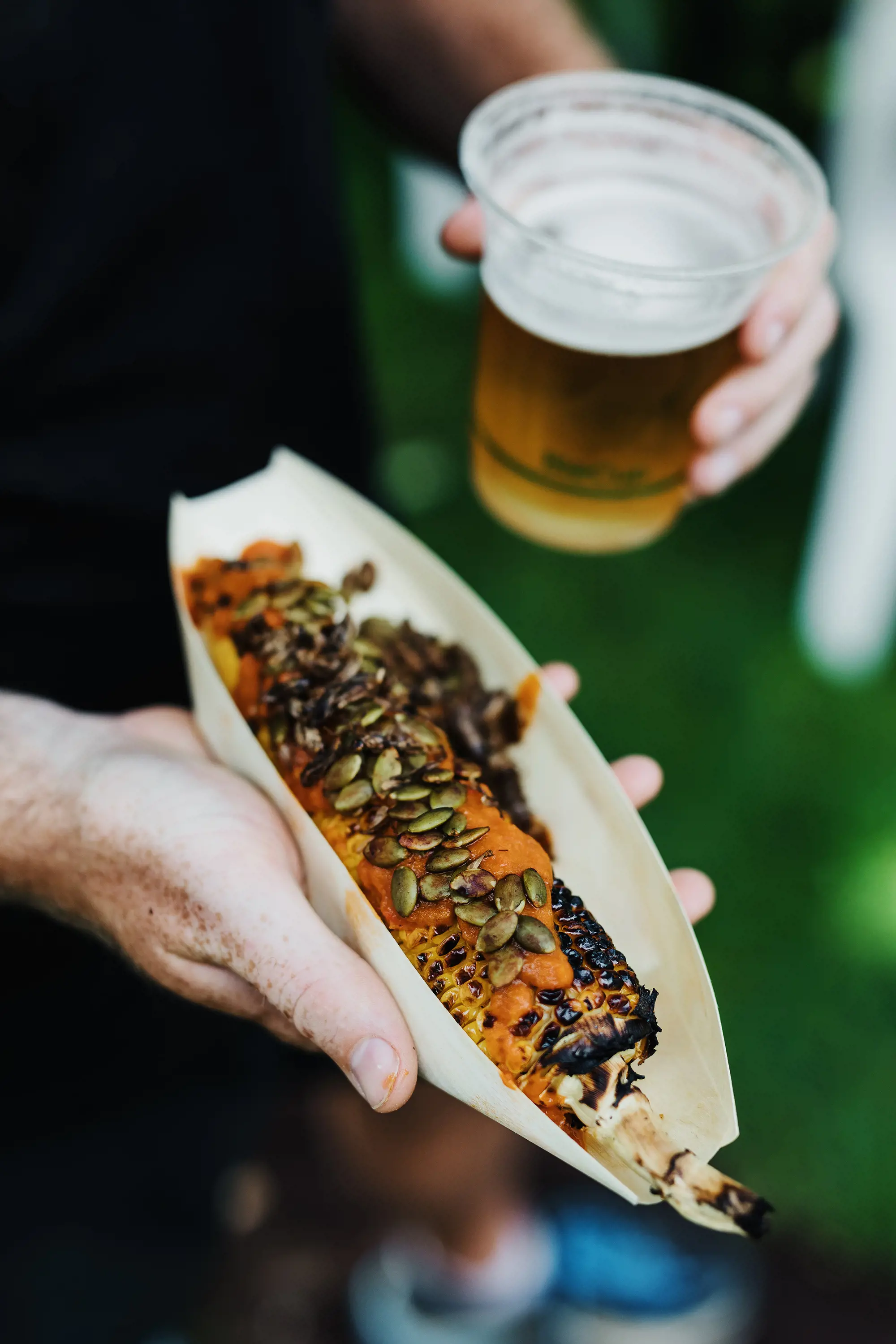 A person's hands holding a plastic cup of beer, and a small food basket of a charred corn cob, topped with orange puree and toasted seeds.