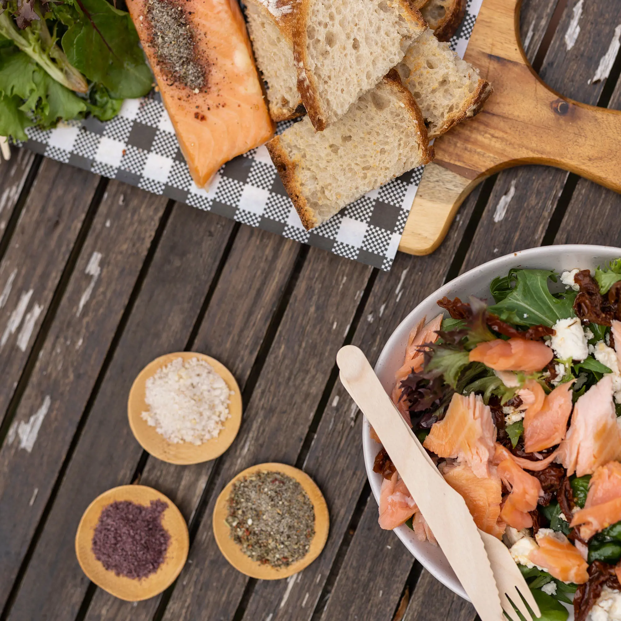 A top-down view of a wooden picnic table with plates of bread, salmon and salad artfully arranged.