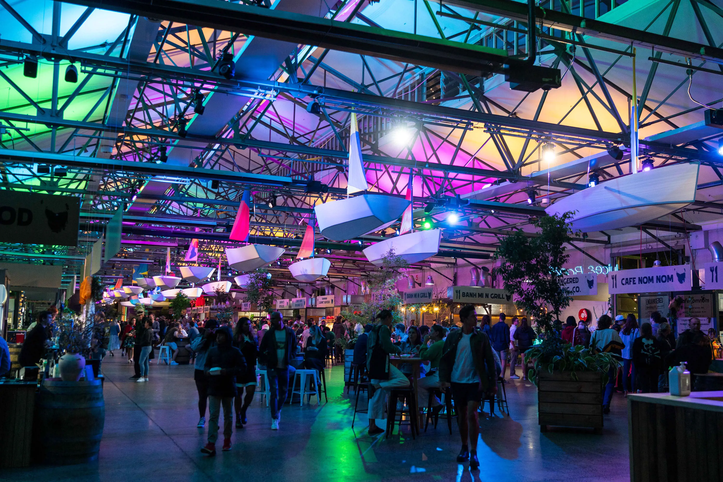 The interior of a large warehouse, with the roof panels lit up in lots of colours. People browse food stalls or sit at bar tables dotted throughout. 