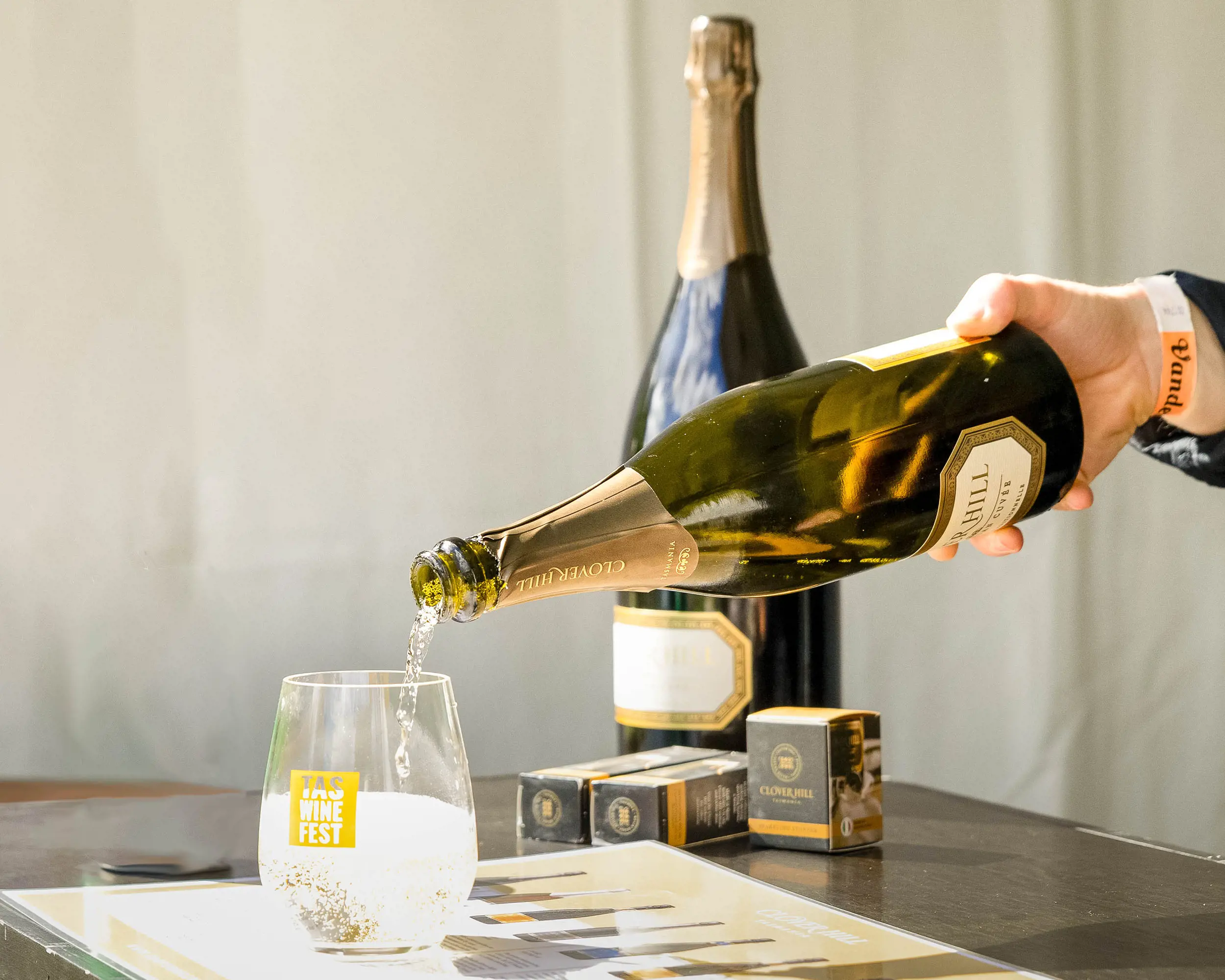 A person's hand holding a champagne bottle, pouring into a reusable clear plastic cup with a logo saying 'Tas Wine Fest' on it.