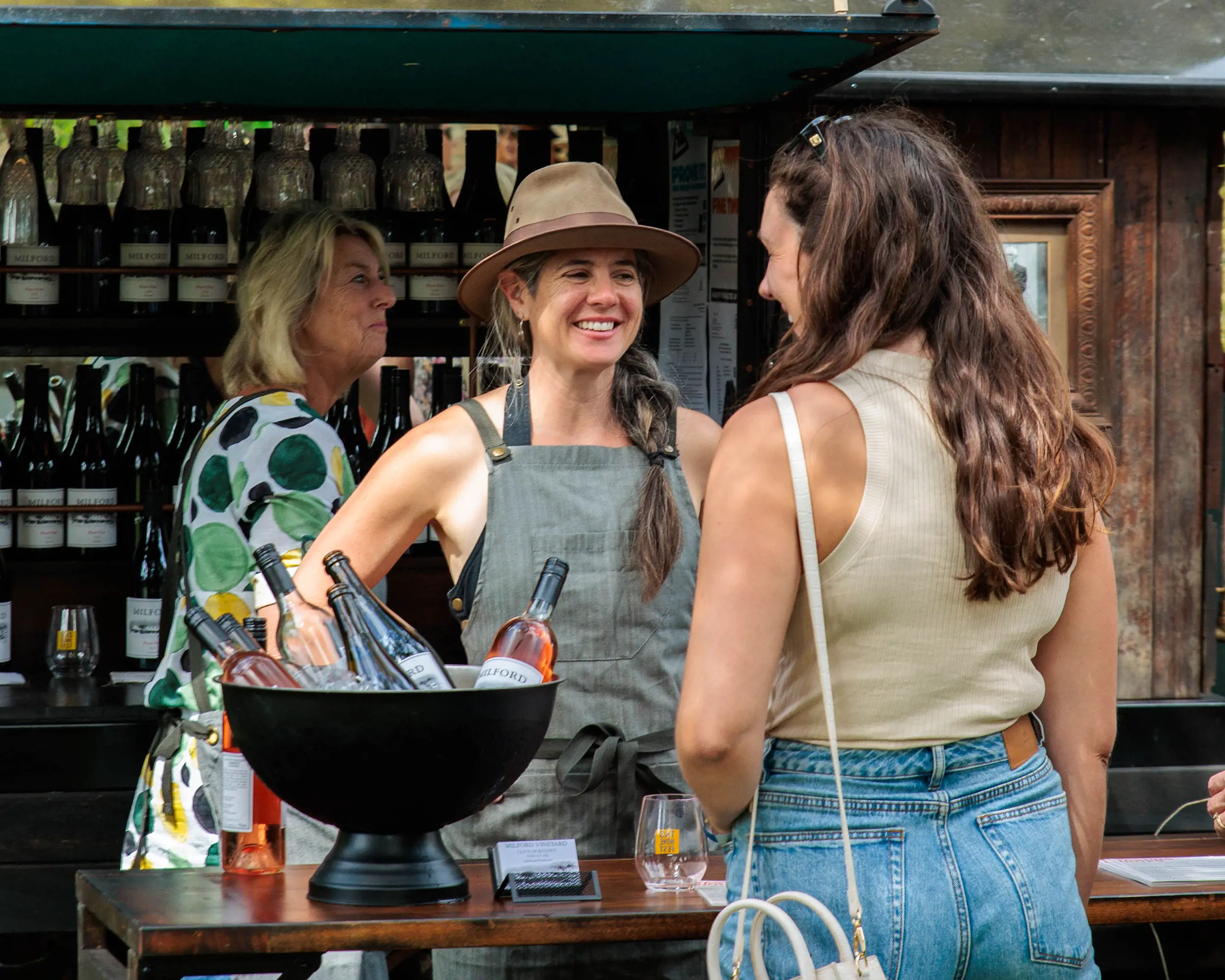 A customer chats to two people working at a festival wine stand, with bottles lined up on shelves behind them and an icebucket with bottles on the counter.