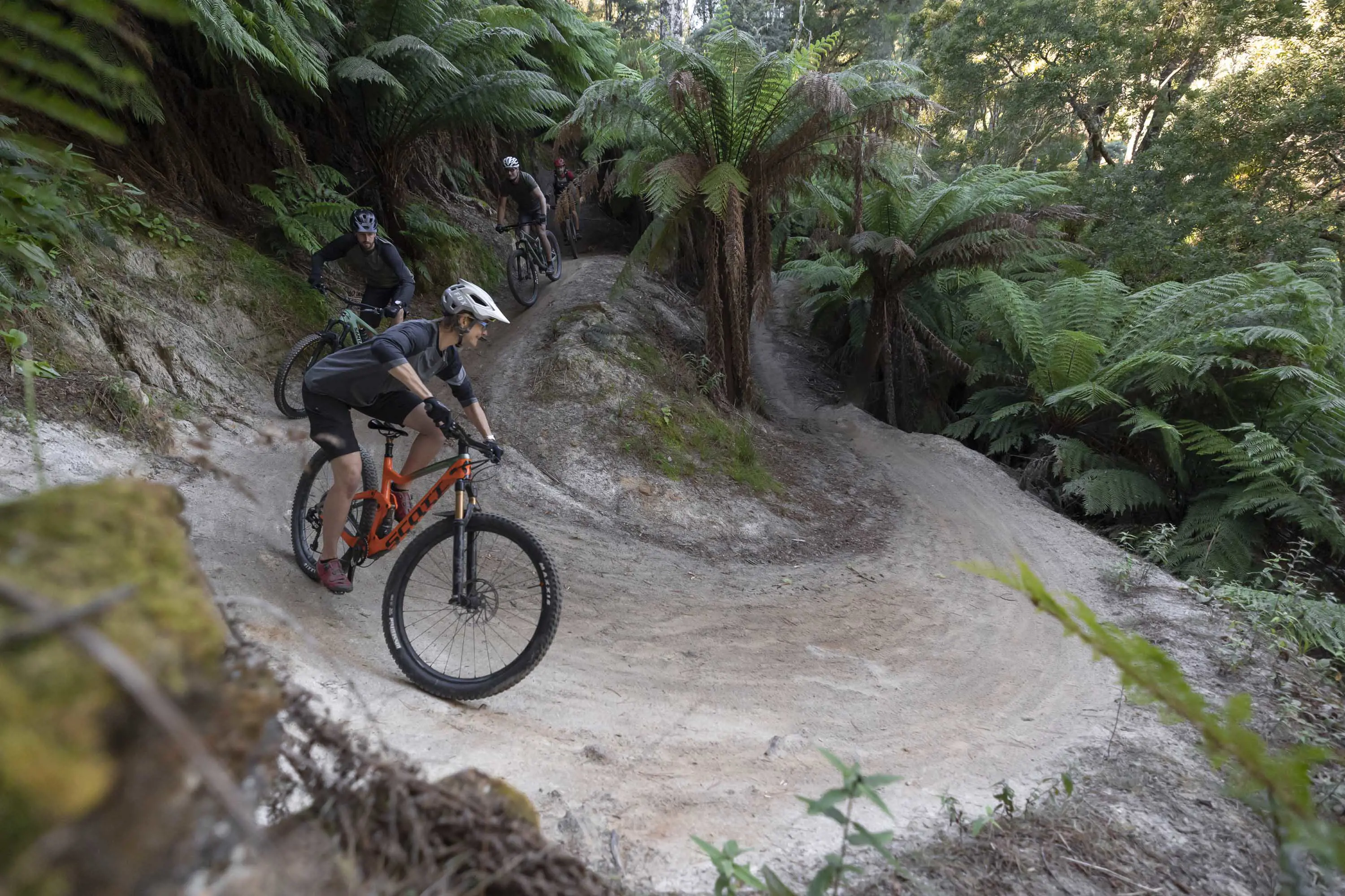 A young rider takes a gravel corner that winds around and through large ferns and forest.
