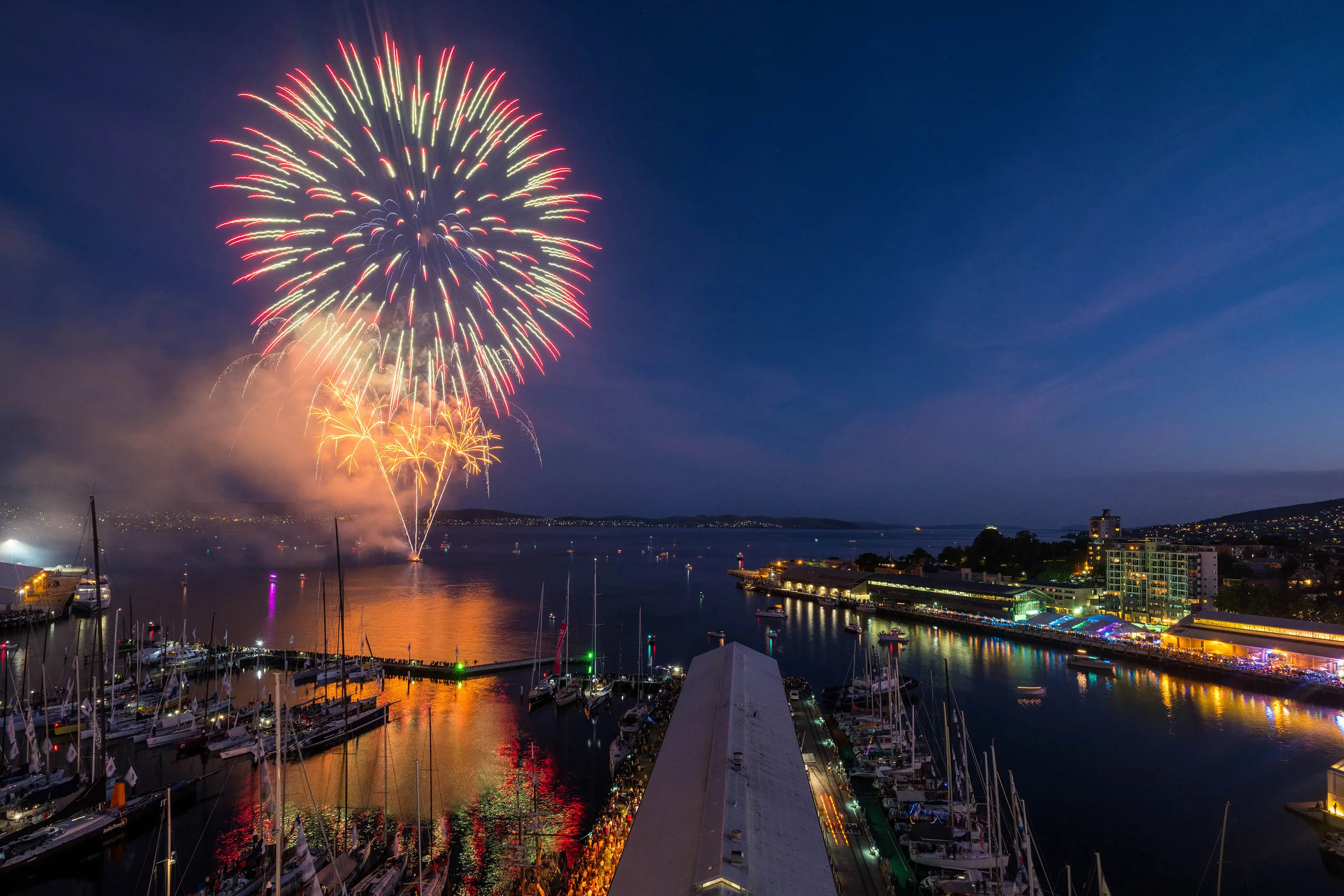 A large fireworks display at the waterfront of Hobart is reflected in the calm waters of a river.