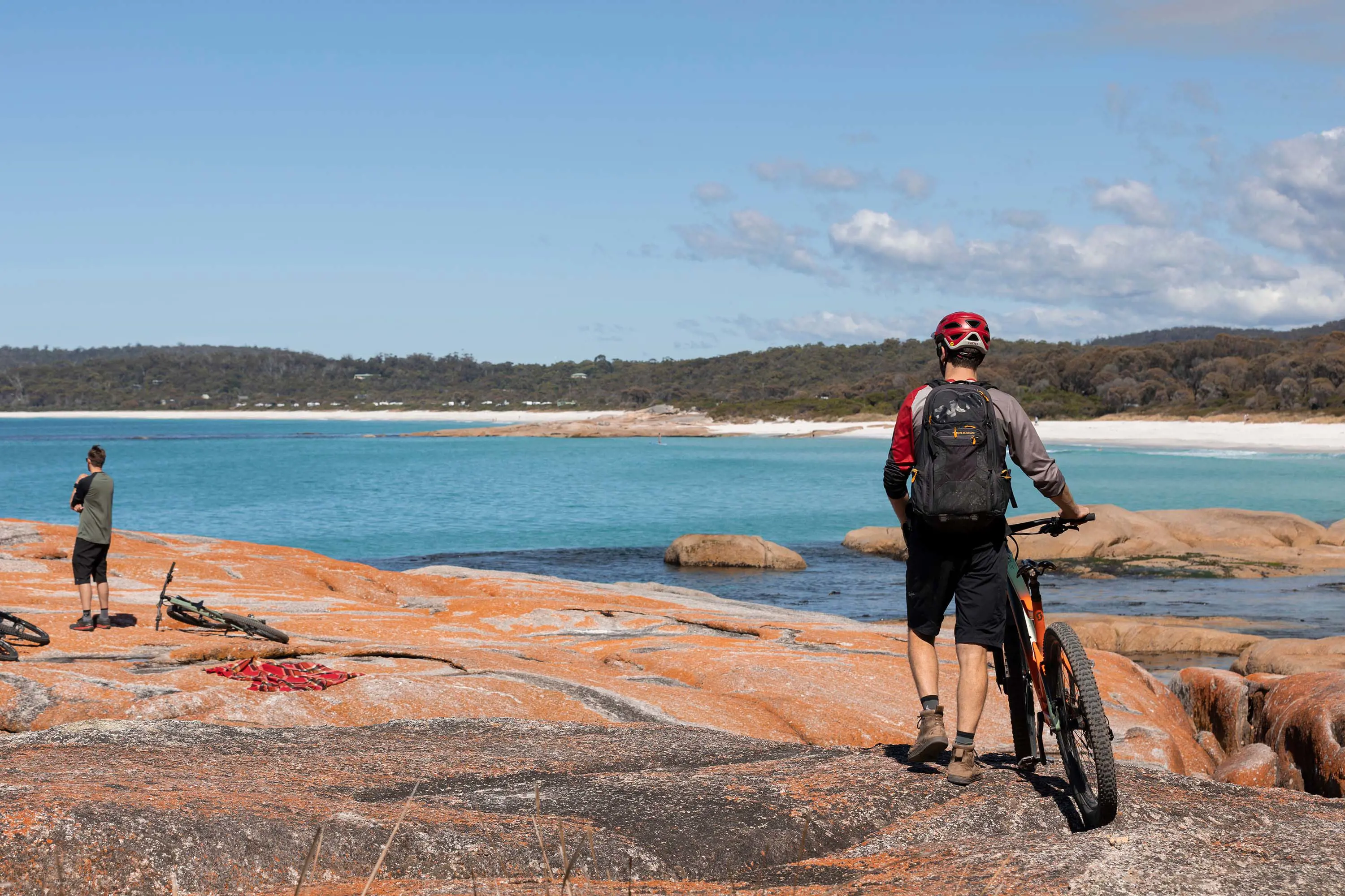 A mountain biker walks his bike across wide, flat rocks next to the clear blue waters of a beach.