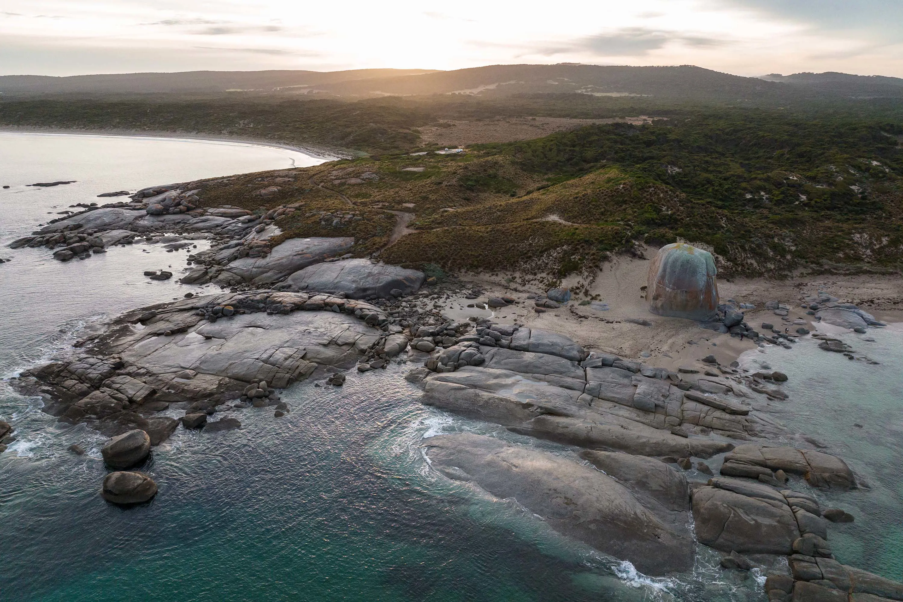 An aerial view of a beach with large rocks reaching out into the water. The sun shines just above the horizon.