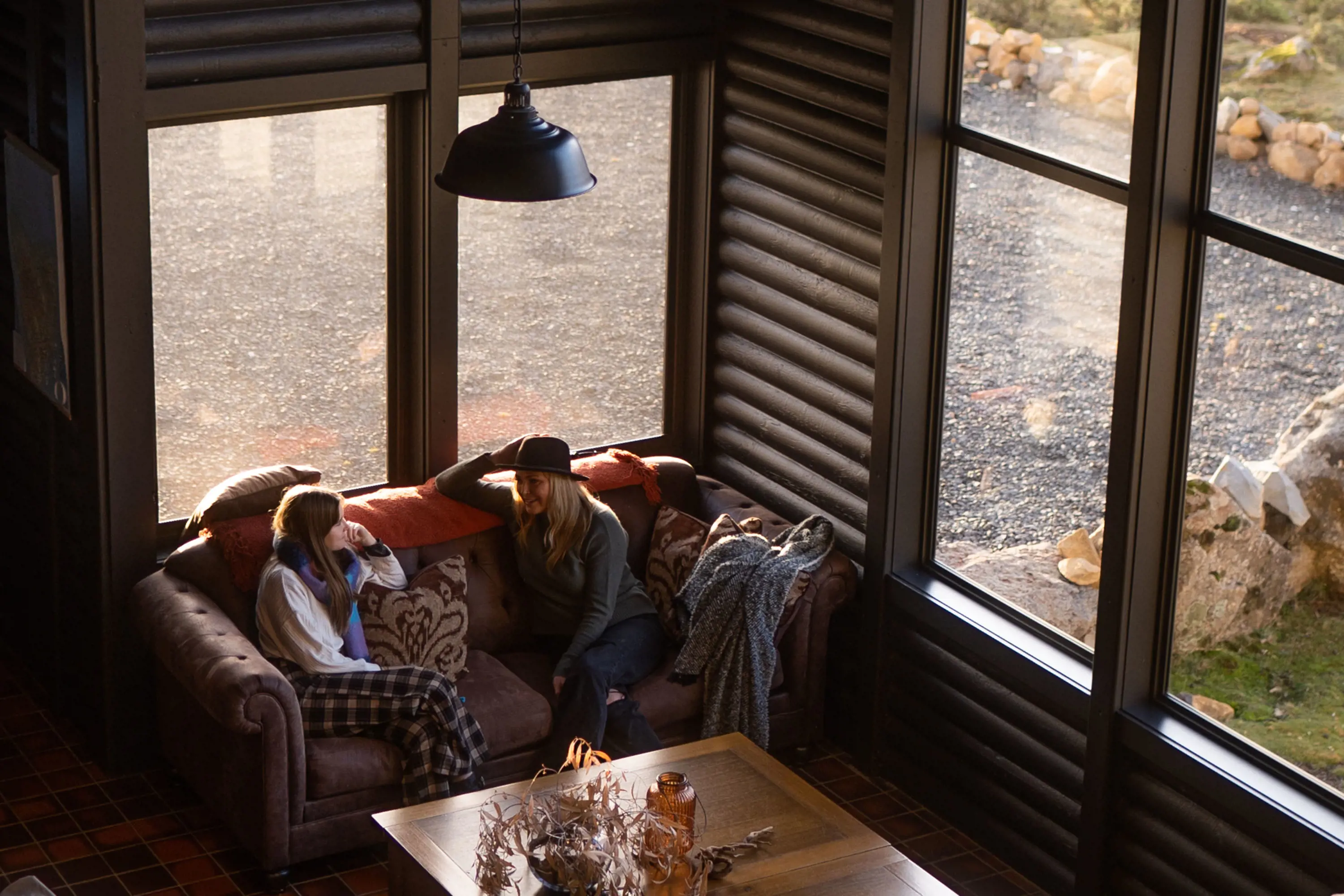 Two people sit on a cozy couch tucked into the corner of a loungeroom, with corrugated iron walls and large windows.