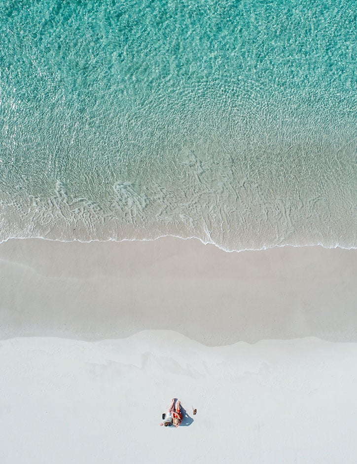 A striking aerial photo of the waterline at a beach. The water is crystal clear aqua blue, and two small figures relax on the white sand.