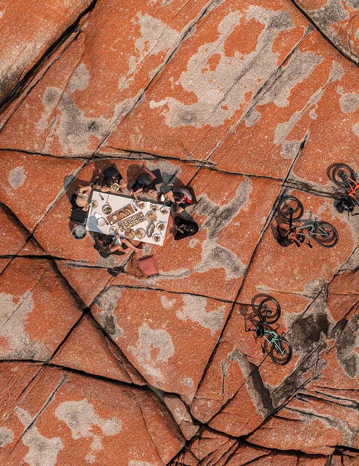 A top-down view of a group of people having a picnic on some vividly orange coloured rocks. Nearby, a few mountain bikes are laid on their sides.