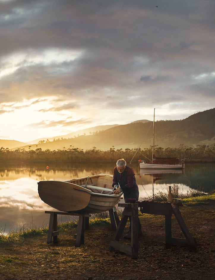 By the banks of a still, picturesque river, a man works on a wooden dinghy propped up on two sawhorses.