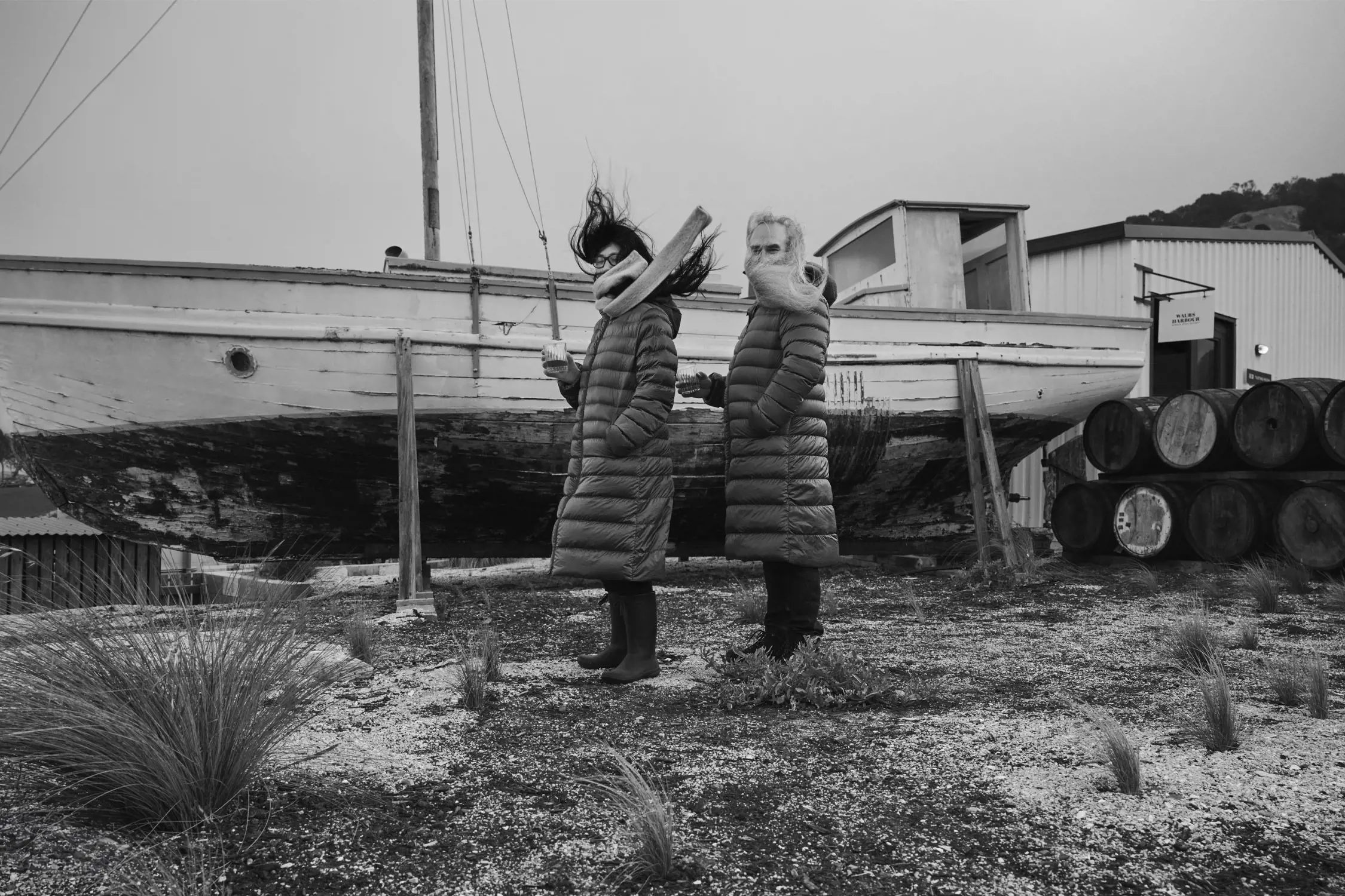 A black and white image of two people in knee length puffer cotas, scarves and gumboots. They are standing next to a boat and barrels. Their hair wips round them in the strong wind. 