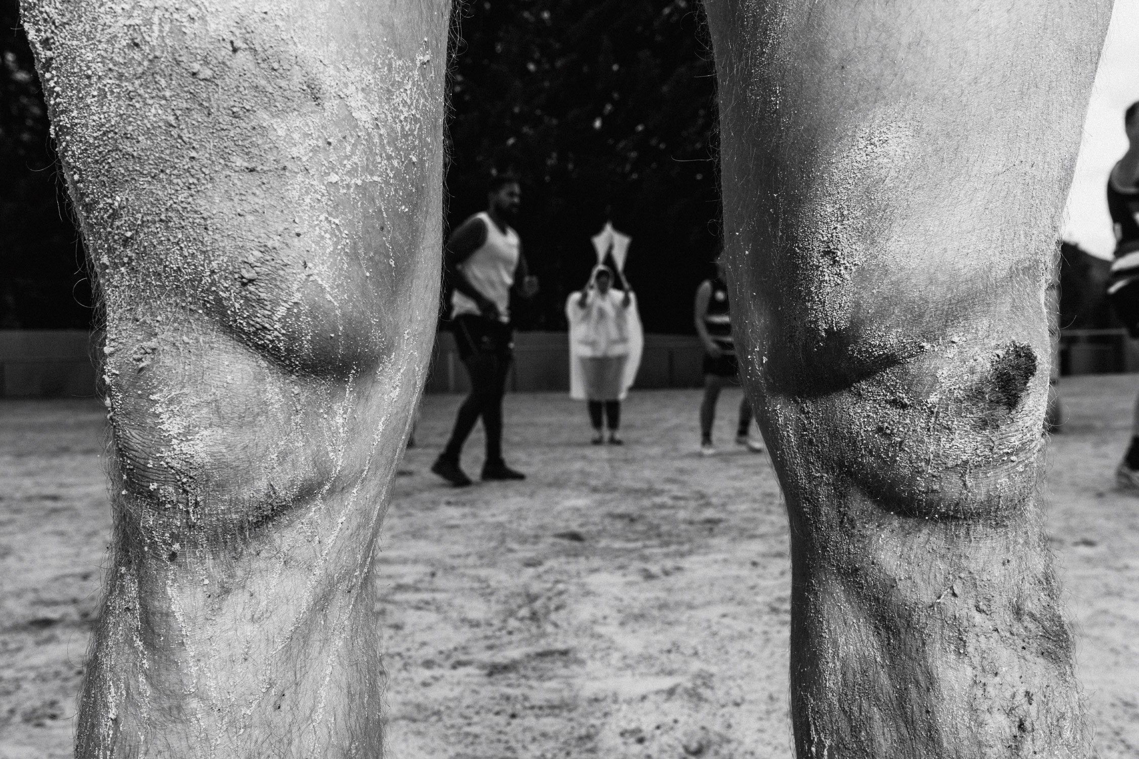 A close-up of human knees caked with dried mud. In the background, a group of people blurred in motion, in the middle of a football game. 