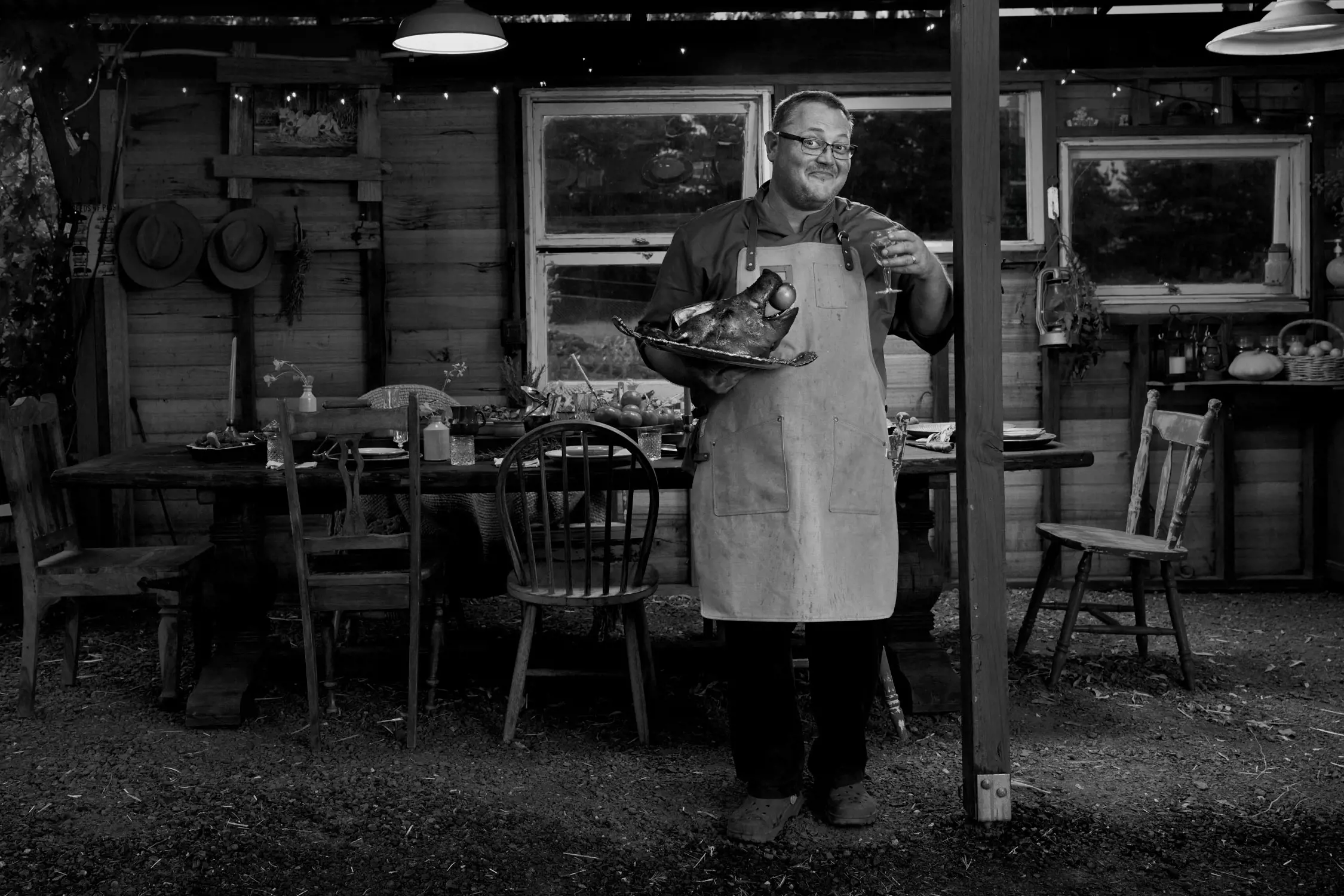 The image depicts a man standing under a rustic outdoor shelter, holding a tray with a roasted pigs head with an apple in it's mouth. He is wearing an apron and smiling warmly. Behind him, there is a wooden table set with dishes and chairs, creating a cozy, communal dining setup. The background includes a wooden structure with windows, shelves, and various items such as hats and kitchen tools, giving the scene a homely, farmhouse atmosphere. Soft lighting and natural surroundings enhance the welcoming vibe.