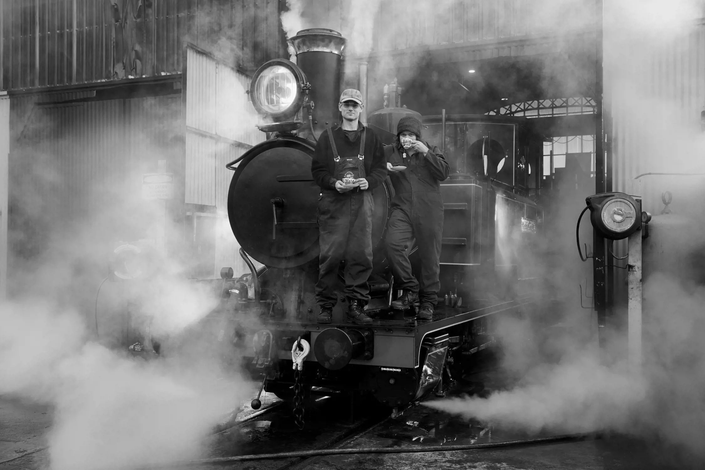 The image features two workers standing on the front of a vintage steam locomotive. Both are dressed in overalls and workwear, with one wearing a cap, holding a teacup and sacuer, and the other wearing a beanie drinking from a teacup. The scene is enveloped in thick steam, creating a dramatic and industrial atmosphere. The locomotive is positioned outside a train depot or workshop with large metal doors and visible structural details in the background. The lighting highlights the machinery and the workers, emphasizing the nostalgic and mechanical essence of the setting.