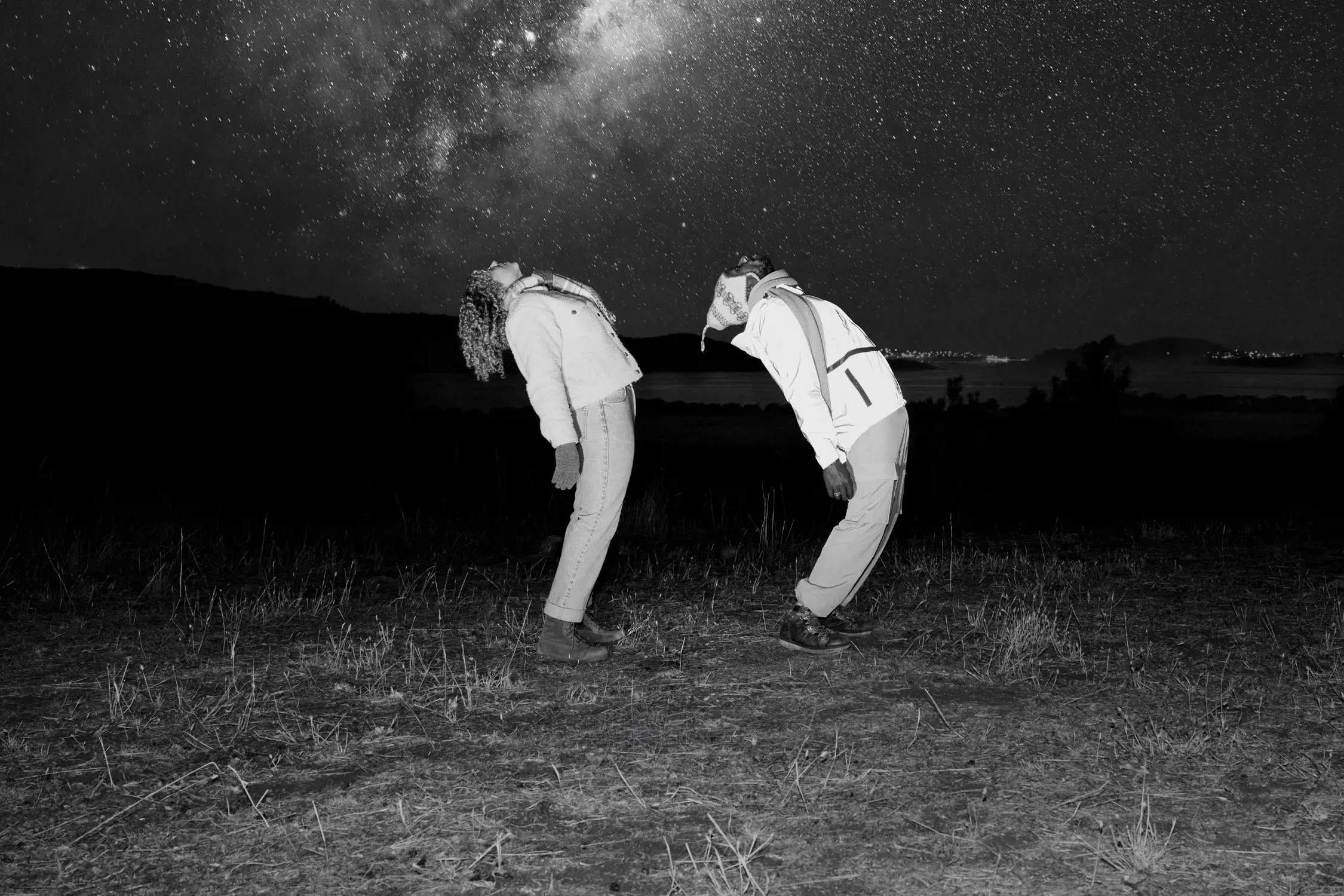 The image depicts two people dressed in jackets and winter clothing bending backward in a synchronized pose under a star-filled night sky. The Milky Way galaxy is prominently visible, creating a striking backdrop. The scene appears to be in an open field with faint outlines of distant hills and possibly a body of water in the background. The image is in black and white, highlighting the contrast between the glowing stars and the dark silhouettes of the landscape.