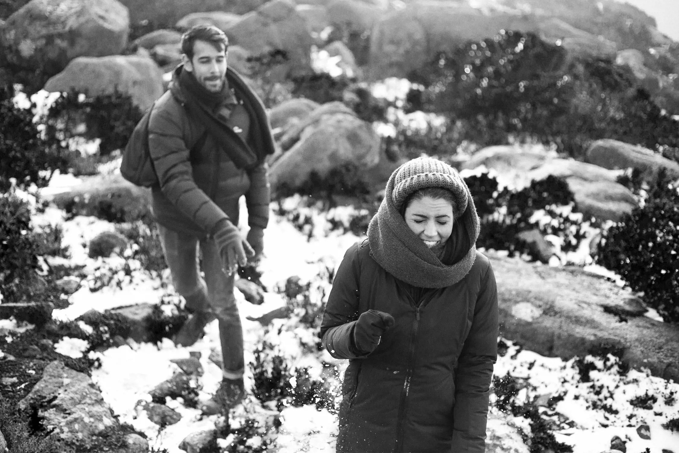 The image shows two people enjoying a snowy outdoor scene. In the foreground, a woman dressed warmly in a jacket, scarf, and knit beanie appears to be laughing as snow flies near her. Behind her, a man wearing a padded jacket and scarf, carrying a backpack, is mid-motion, possibly tossing snow. The ground is covered with patches of snow and scattered rocks, surrounded by shrubs and boulders. The background is a natural, rugged landscape with a soft, wintry atmosphere