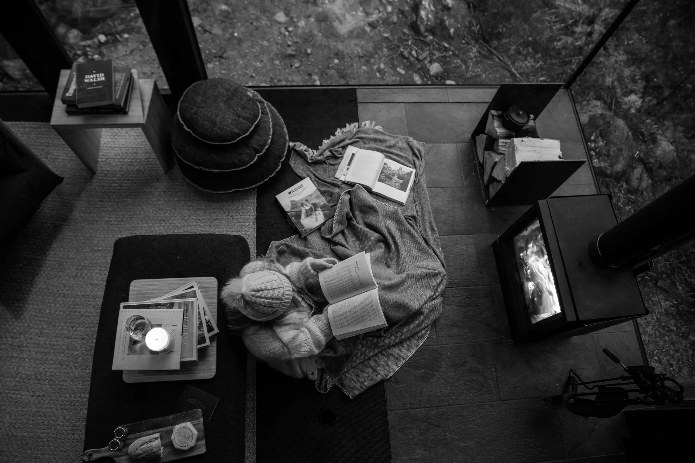 A cozy indoor scene viewed from above, showing a person sitting on a soft rug near a fireplace, wrapped in a blanket and wearing a knitted beanie. They are reading an open book, with additional books and magazines scattered around. Nearby, a wooden table holds a lit candle, a stack of books, and a small charcuterie board with bread and cheese. The area is surrounded by large glass windows offering a view of the outdoors, with a mix of natural elements like rocks and soil visible. The space exudes warmth and tranquility, enhanced by the glowing fireplace.