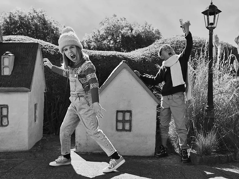 Two kids dressed warmly in scarves and beanies roar and playfully stomp through the streets of a miniature village.