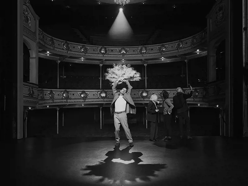 A man dramatically poses with beautiful burlesque fans under the spotlight on a historic theatre stage.