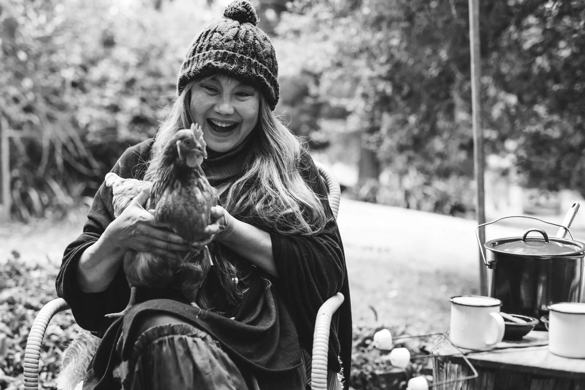 A woman sitting outdoors laughs heartily while holding a chicken in her arms. She wears a warm knit hat and layered clothing. Nearby, a rustic wooden table holds enamel mugs and a cooking pot, adding to the cozy and lively outdoor atmosphere