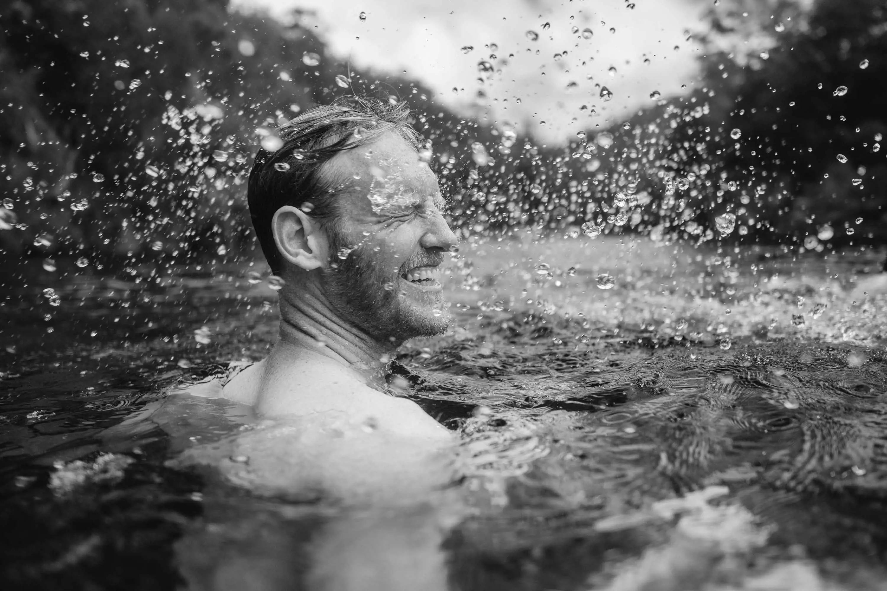 A young man with a beard up to his shoulders in water. He's shaking his head and water droplets fly through the air around him.