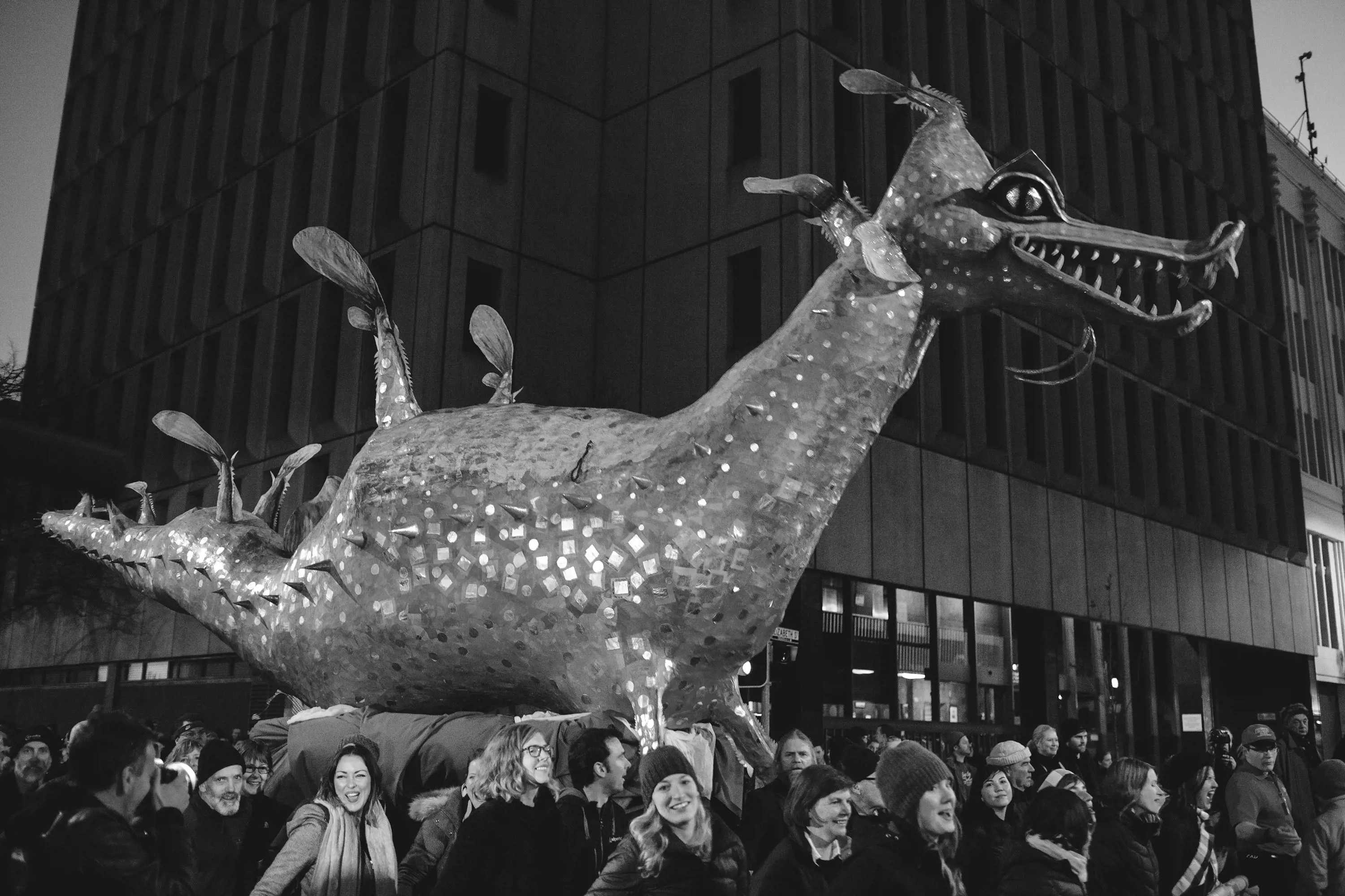 A black and white image of a large stylised dragon-like creature, carried on a float surrounded by people in beanies, scarves and jackets.