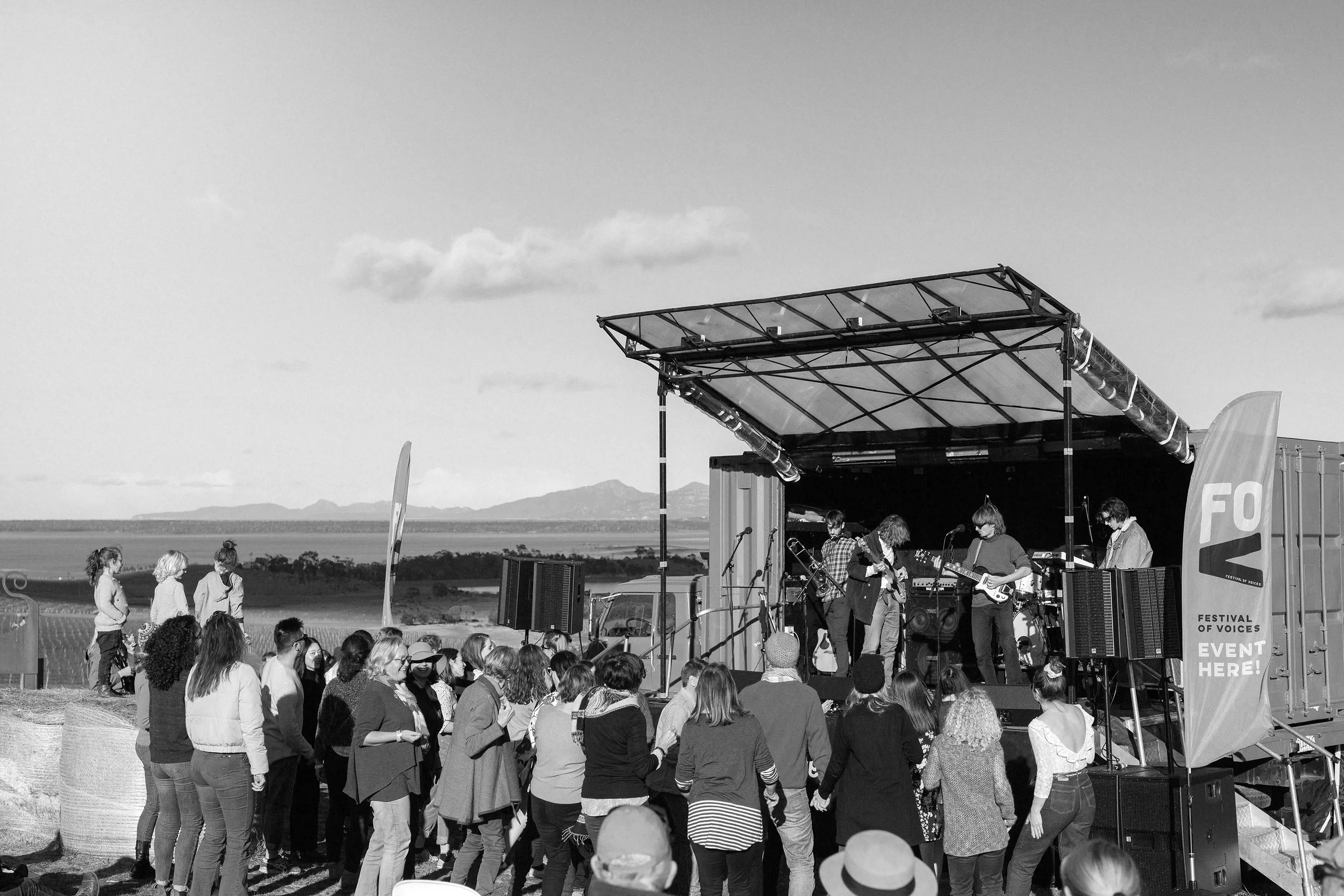 A black and white image of an outdoor pop-up stage, with a band playing and people watching and dancing in front.