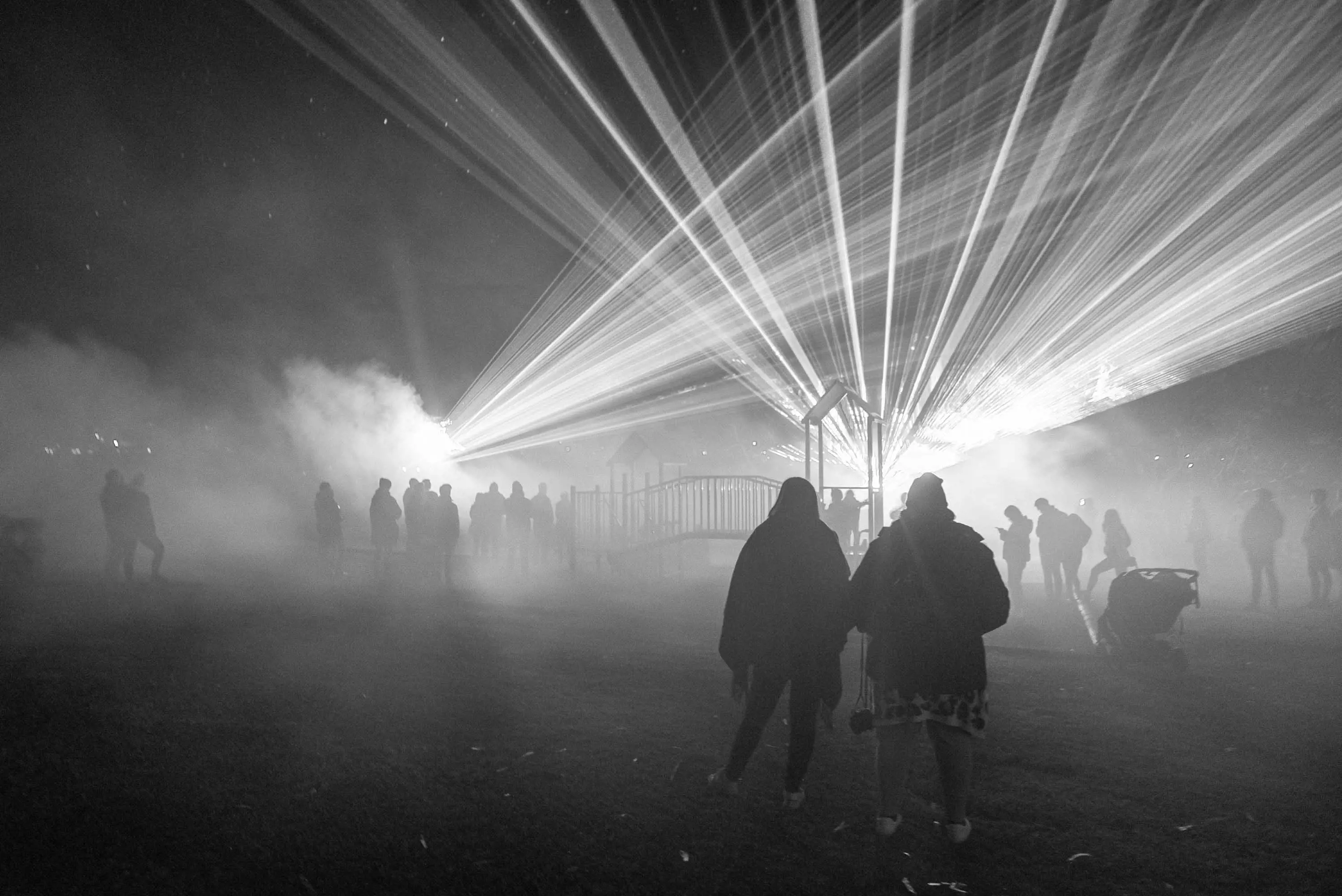 People walk through a dark, smoke filled park. Lasers shine through through and illuminate the smoke and the crowd.