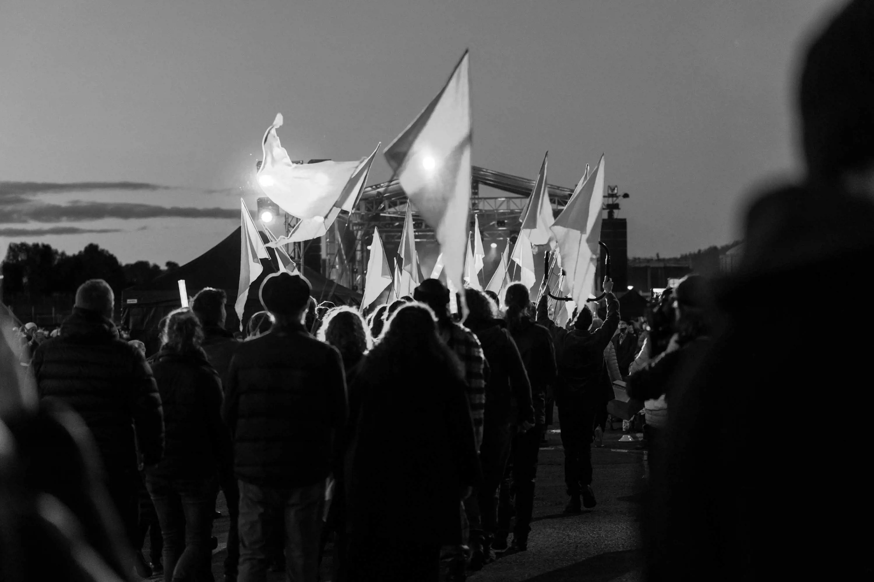A large procession of people carrying flags walk in a long, sprawling line towards a performance stage in the background.
